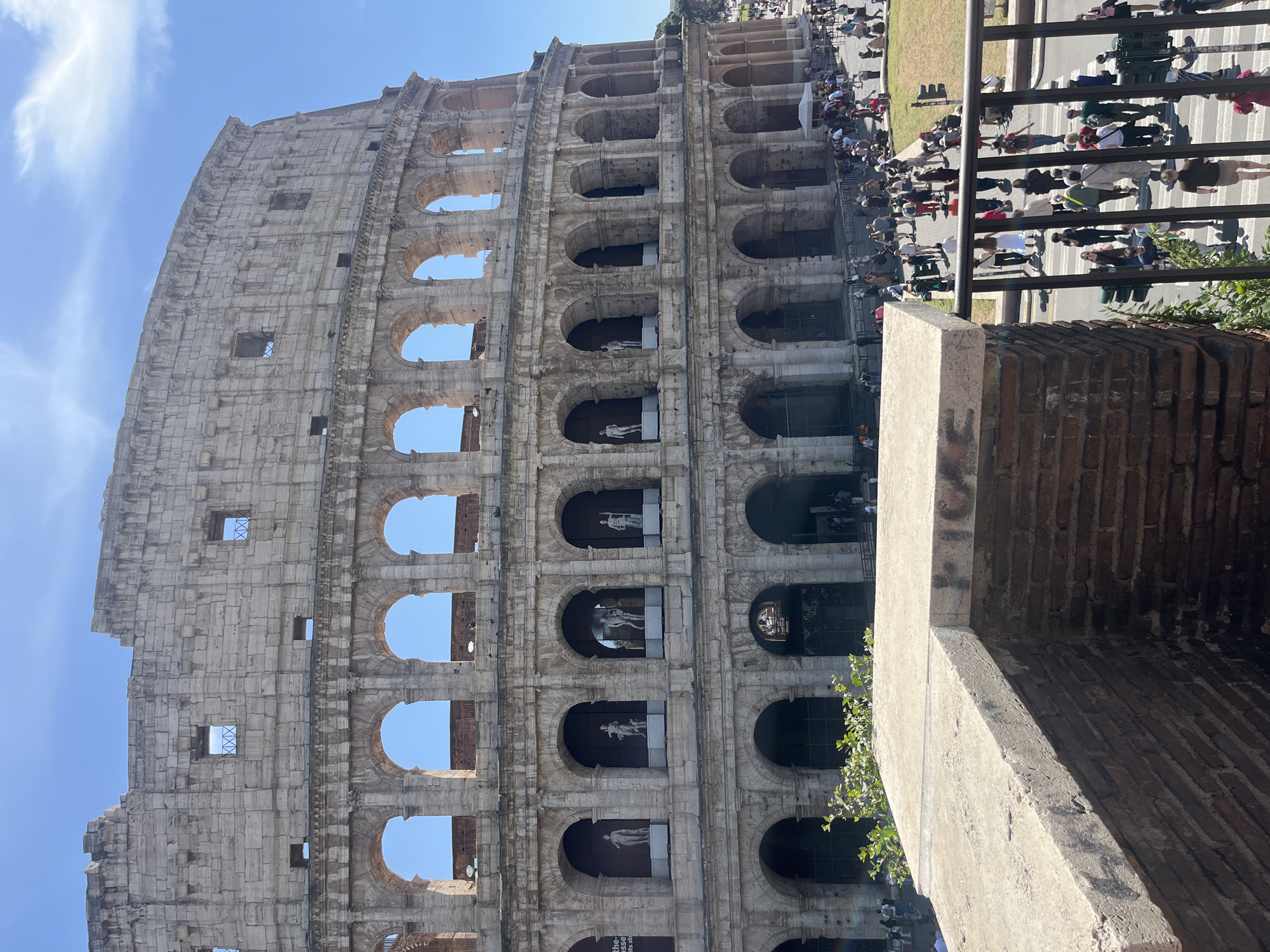 The image captures the Colosseum in Rome, Italy, viewed from an elevated perspective. The Colosseum's multi-tiered structure, composed of stone arches, is prominently displayed. Statues are visible within some of the arches. A crowd of people is observed walking along a street beside the Colosseum, separated by a black metal fence. The weather appears sunny, with a clear blue sky above the ancient structure. In the foreground, a stone wall is partially visible.