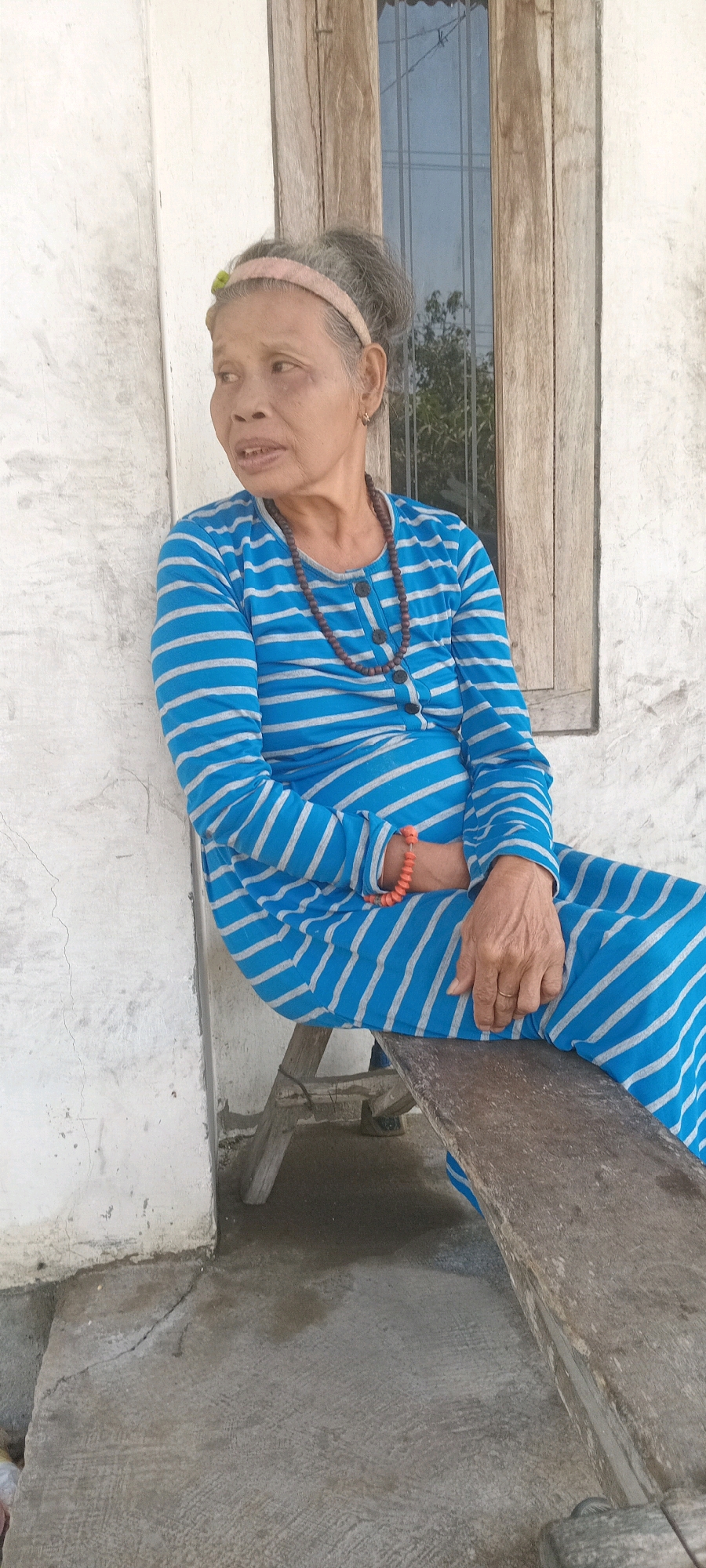 An elderly Asian woman with graying hair tied up and a pink headband sits on a rustic wooden bench outside a building. She is wearing a long-sleeved blue and white striped dress, a beaded necklace, and an orange bracelet on her left wrist. Her left hand rests on her lap, while her right hand is on her right leg, revealing a ring on her index finger. Behind her is a white, textured wall and a wooden-framed window with reflective glass. The scene appears to be located in Kedukbembem, Indonesia, based on the provided context. The ground is concrete, with visible cracks and some dampness.