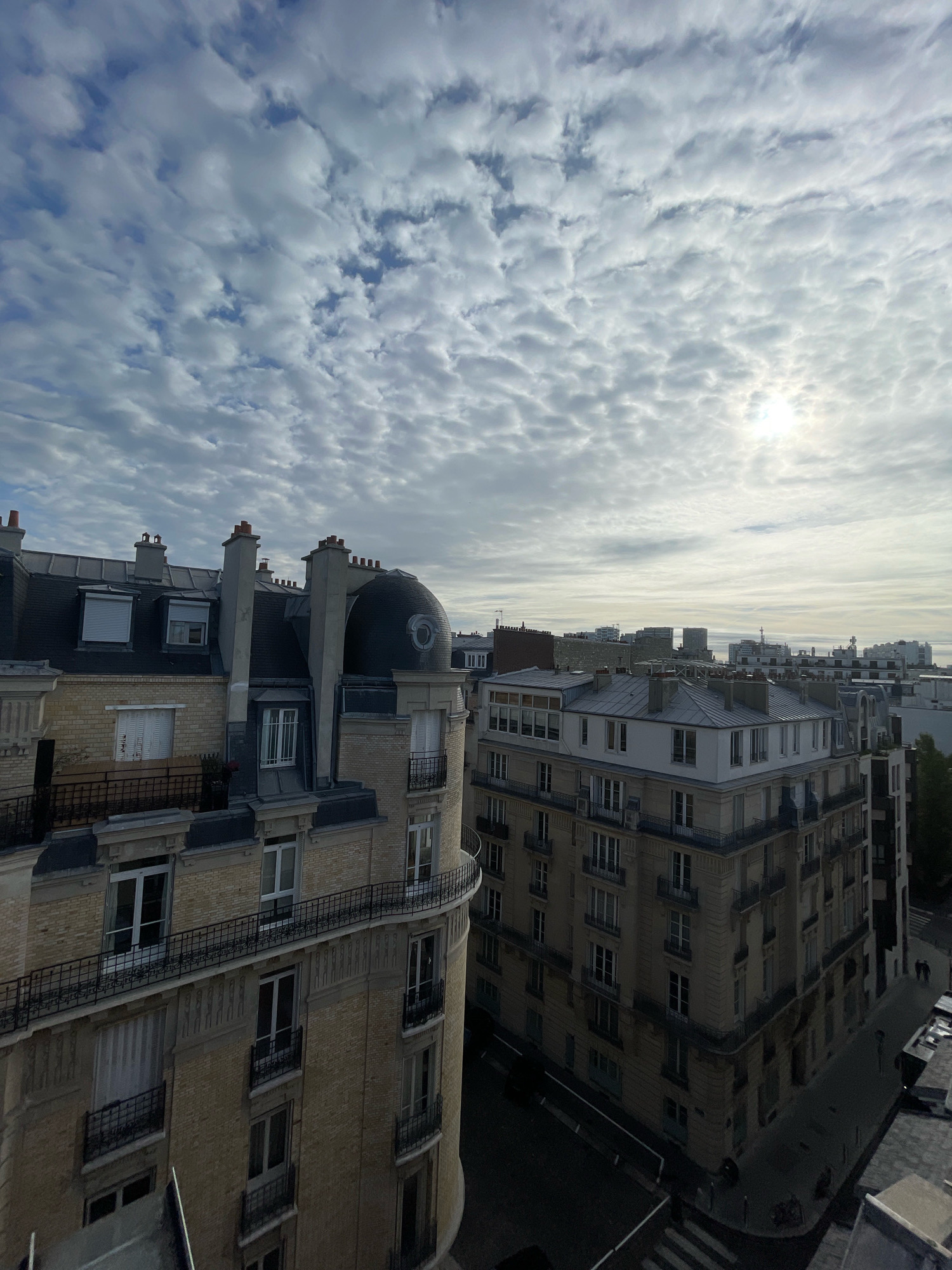 The image is an aerial view of Parisian rooftops and city skyline under a cloudy sky. In the foreground, a light-colored, multi-story building with a curved facade is visible, featuring a dark, dome-shaped roof structure and several chimneys. The building has numerous windows, some with shutters, and ornate balconies with black metal railings. Adjacent to it is another similar building, also with light-colored brickwork, pitched roofs, and chimneys. The middle ground shows a dense urban landscape with more rooftops and modern buildings in the distance. The sky is filled with patchy, white clouds, with the sun partially obscured, creating a diffused light. A few streetlights and trees are visible at street level. Two figures are seen walking on a street in the lower right portion of the image.