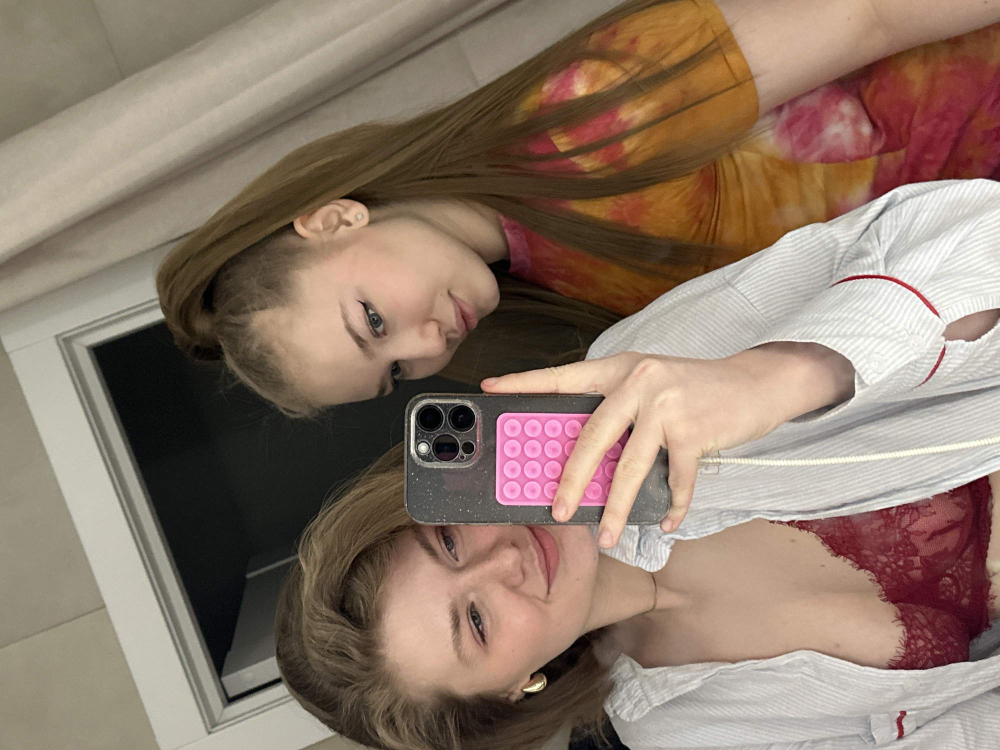 Two women are in the image, taken from a close, elevated angle as a selfie. The woman in the foreground is holding the phone; she has blonde hair, is wearing gold earrings, a white and blue striped top, and a red lace bra is visible underneath. The second woman has long brown hair and is wearing a tie-dye orange and pink top. The background consists of a white dresser or cabinet and a beige curtain. The image location is unknown, and no evidence suggests it is in Kazan, Russia.