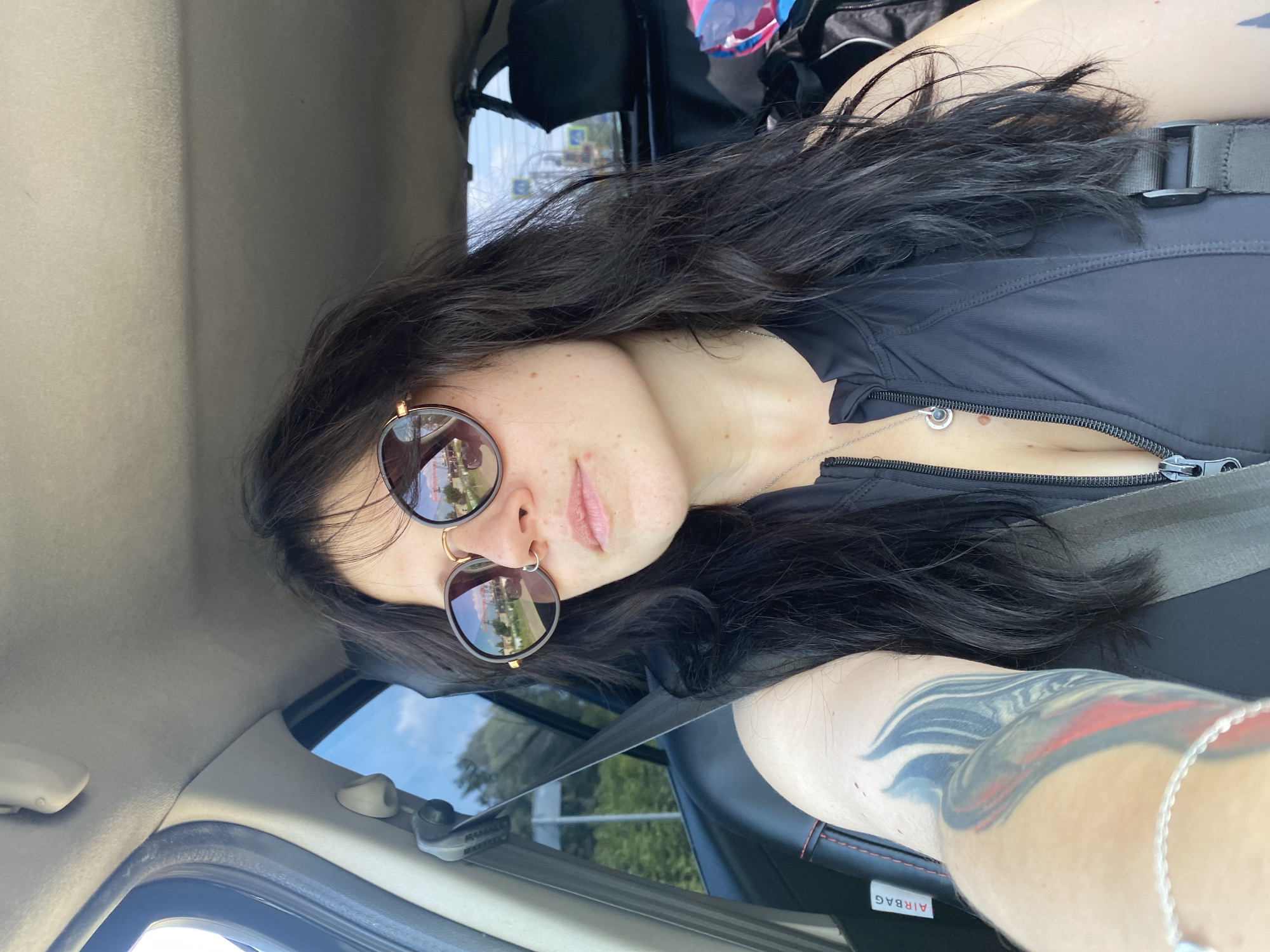 A woman with long, dark hair and sunglasses is taking a selfie while sitting in the driver's seat of a car. She is wearing a black top with a zipper. Her left arm, visible in the foreground, has a tattoo. The car interior, including a seatbelt and an "AIRBAG" label, is visible. Outside the car window, trees and a bright sky can be seen. The reflection in her sunglasses shows buildings and a bridge, suggesting an urban or industrial setting. The car appears to be parked or moving slowly on a road.