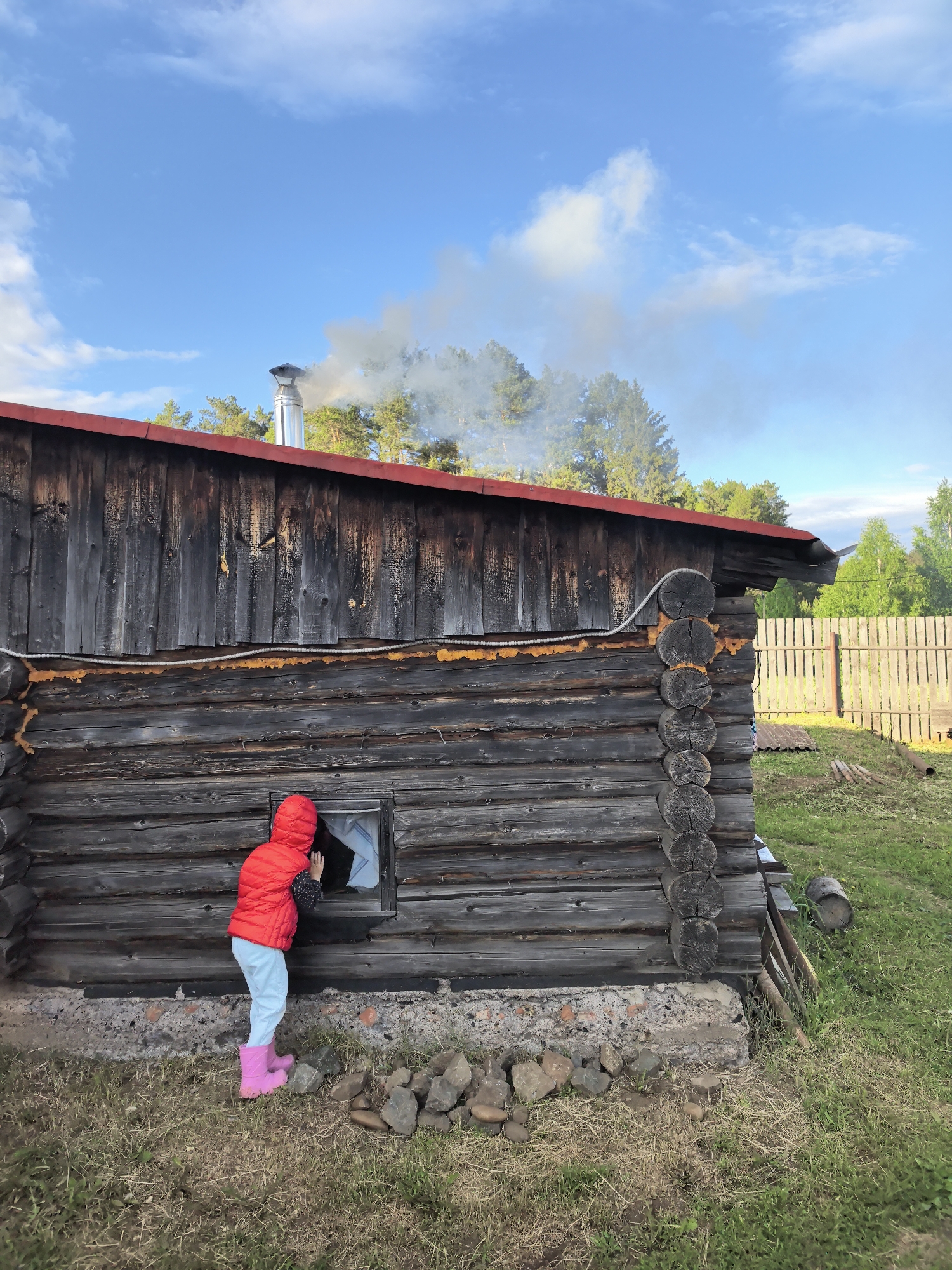 A young person in a red jacket, light blue pants, and pink boots stands outside a rustic wooden cabin, looking into a window. The cabin has a red roof and a metal chimney that is emitting smoke, suggesting a fire is burning inside. The building's foundation appears to be a mix of concrete and rocks. The surrounding environment features green grass and trees, with a wooden fence visible in the background under a partly cloudy sky. The location is Unknown, Unknown.