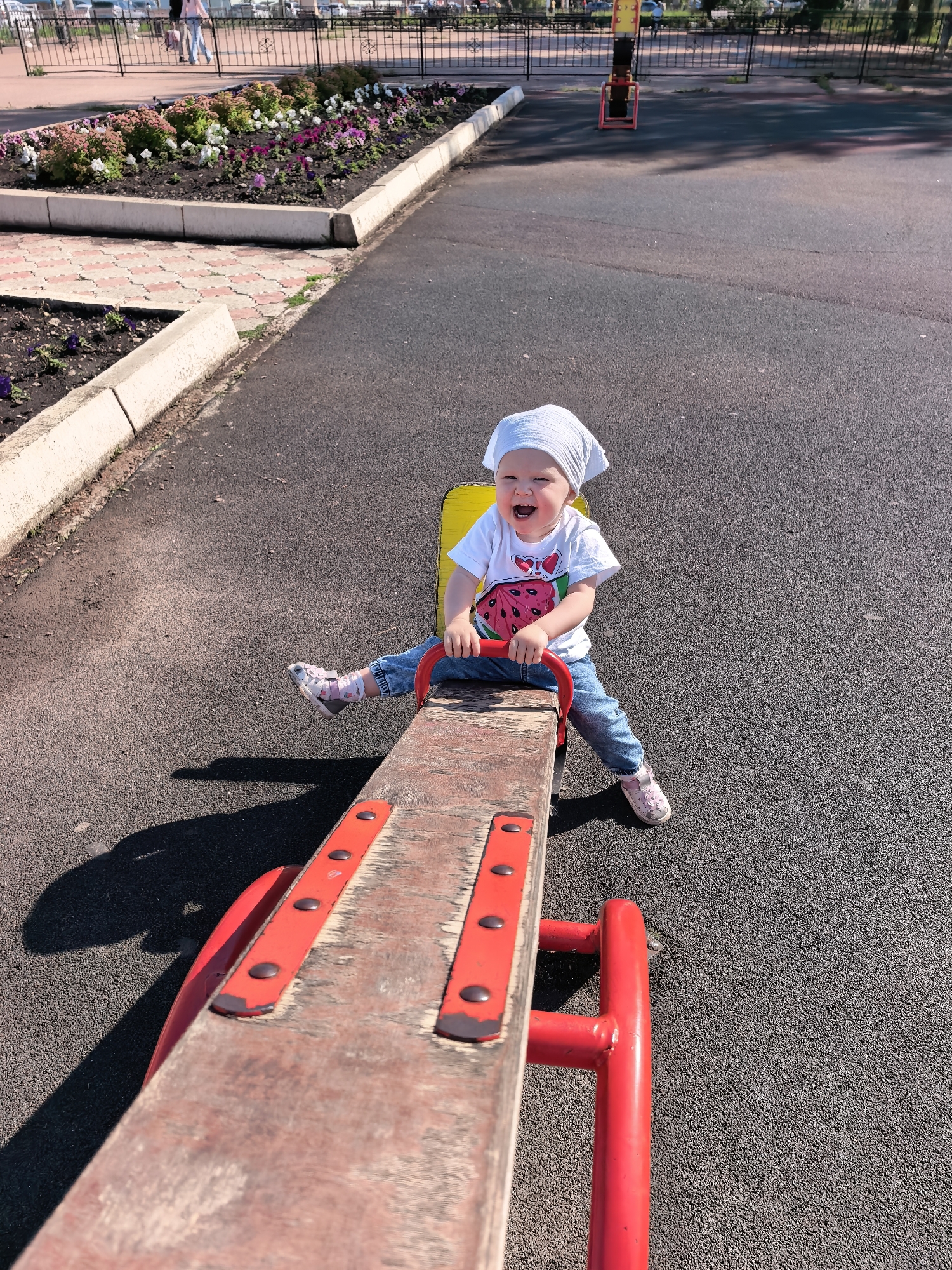 A young child is joyfully riding a seesaw on a playground. The child is wearing a white bandana, a white t-shirt with a watermelon graphic, blue jeans, and pink shoes. The seesaw is made of wood and painted red on the sides. The playground has an asphalt surface and a flower bed with various colorful flowers is visible in the background, bordered by a concrete curb. Beyond the flower bed, a black metal fence encloses a paved area with more playground equipment. In the distance, blurry figures of people can be seen walking. The sun is shining, casting shadows on the ground, indicating it is daytime. The overall scene conveys a happy and playful atmosphere. No visible text is present in the image. The location is Sterlitamak, Russia.