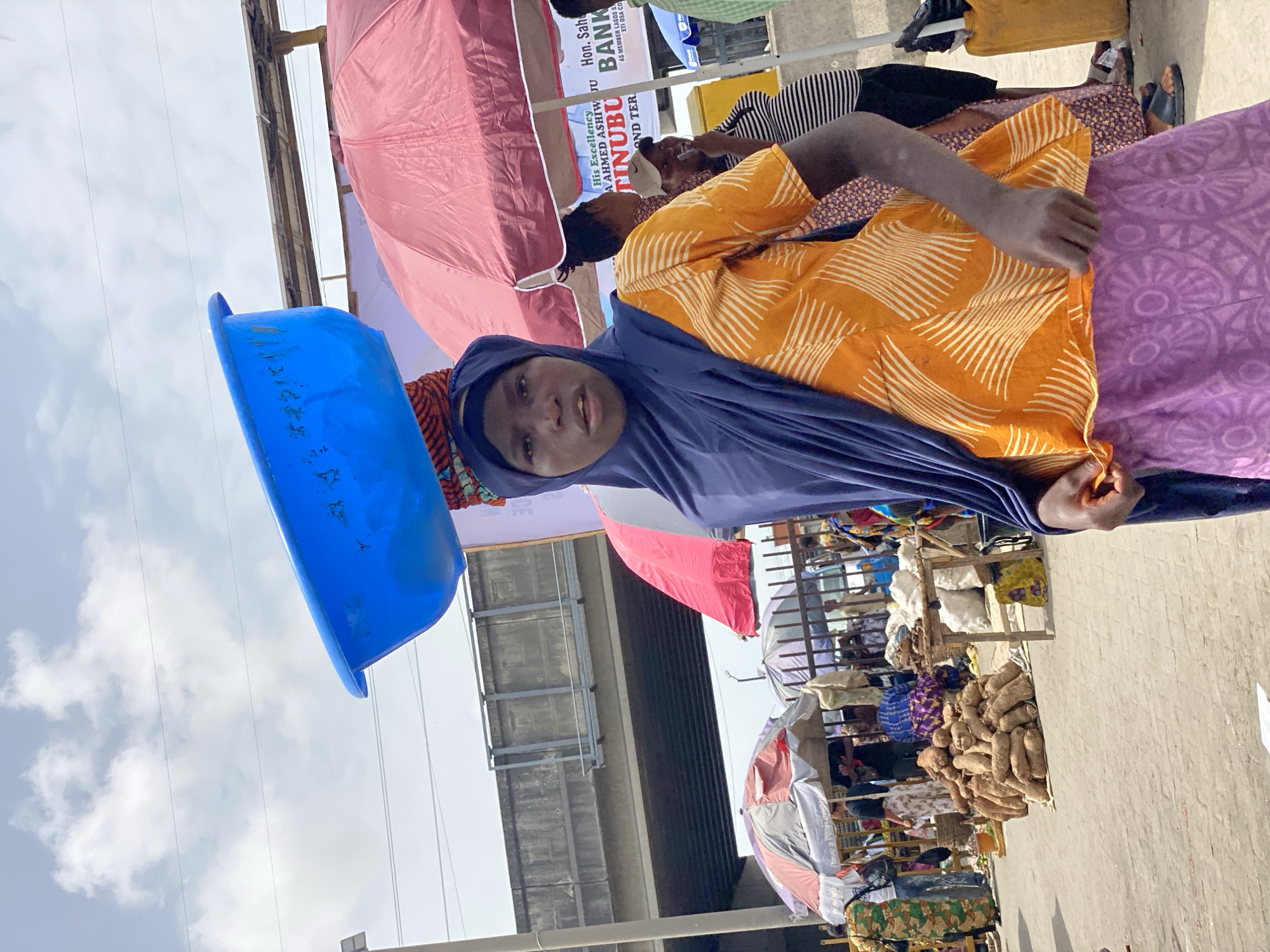 A woman in Lagos, Nigeria, wearing a navy-blue hijab, an orange patterned blouse, and a purple patterned skirt, carries a blue plastic basin on her head. She is positioned at a market. Visible in the background are various vendors with their goods displayed, including yam. Political banners promoting “TINUBU” are seen, along with umbrellas. The scene is under an overpass structure.