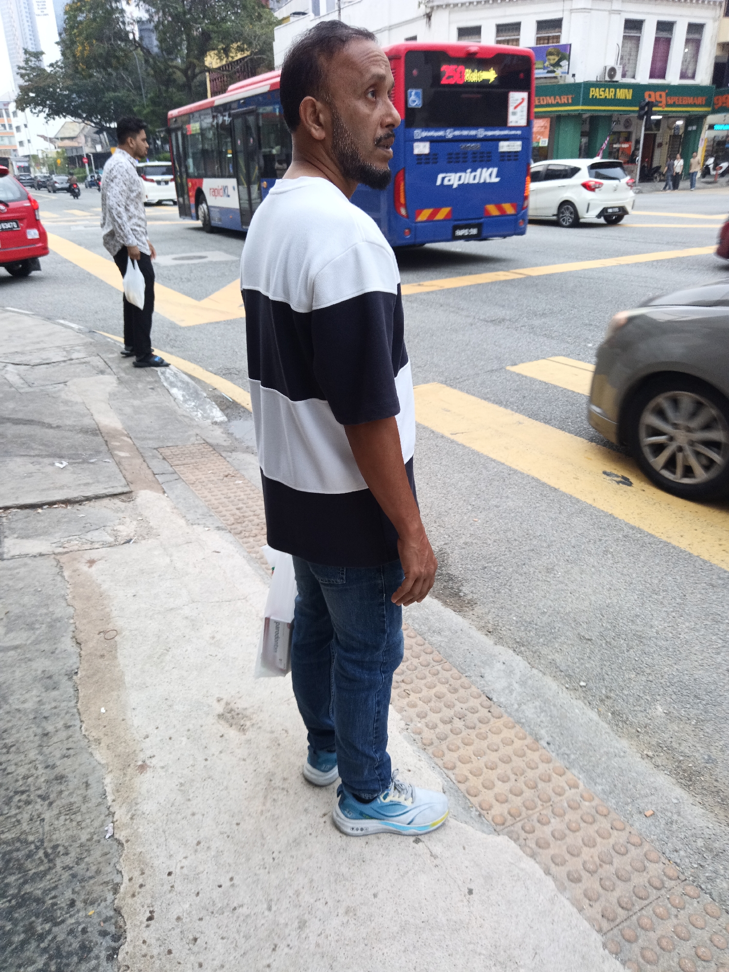 A man stands on a sidewalk next to a street in Kuala Lumpur, Malaysia. He is wearing a black and white horizontally striped t-shirt, blue jeans, and blue and white athletic shoes. He is holding a white plastic bag in his left hand. The man is facing to the right, looking towards the street. In the background, a blue and red bus with "rapidKL" written on it is stopped at a bus stop. A white car is also visible behind the bus. The street has yellow lines marking lanes and a crosswalk. The sidewalk is made of concrete and has textured tactile paving near the curb. Buildings are visible in the background, including shops with signs in Malay.