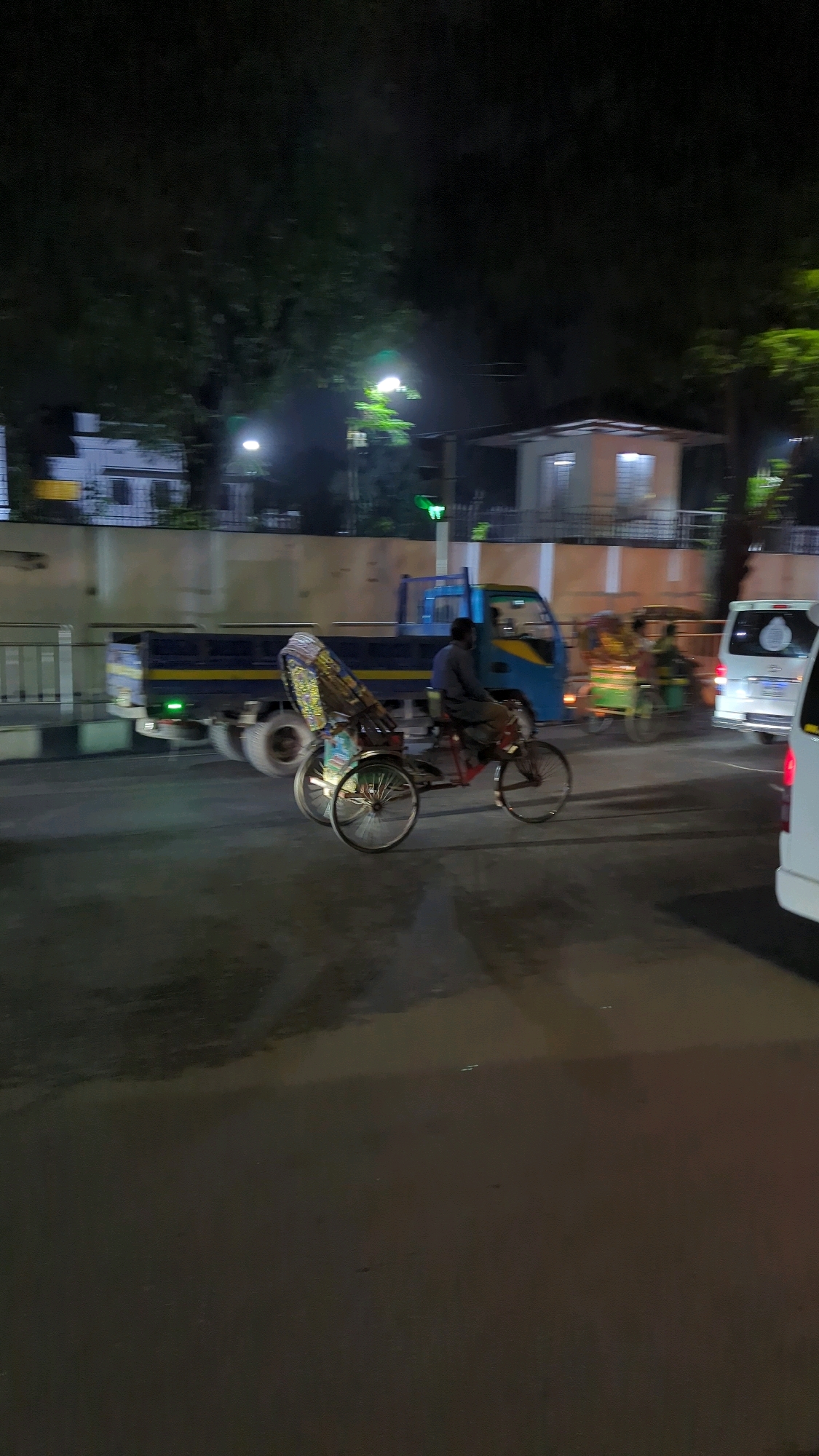 The image depicts a street scene at night in Dhaka, Bangladesh. In the foreground, a cycle rickshaw with a colorful canopy is moving, driven by a man. Behind it, a blue truck is visible, and further in the background, there is a white van and another, blurred rickshaw. The environment appears to be a city street with trees and buildings lining the road, illuminated by streetlights and vehicle headlights. The road surface shows reflections, suggesting it may be wet.