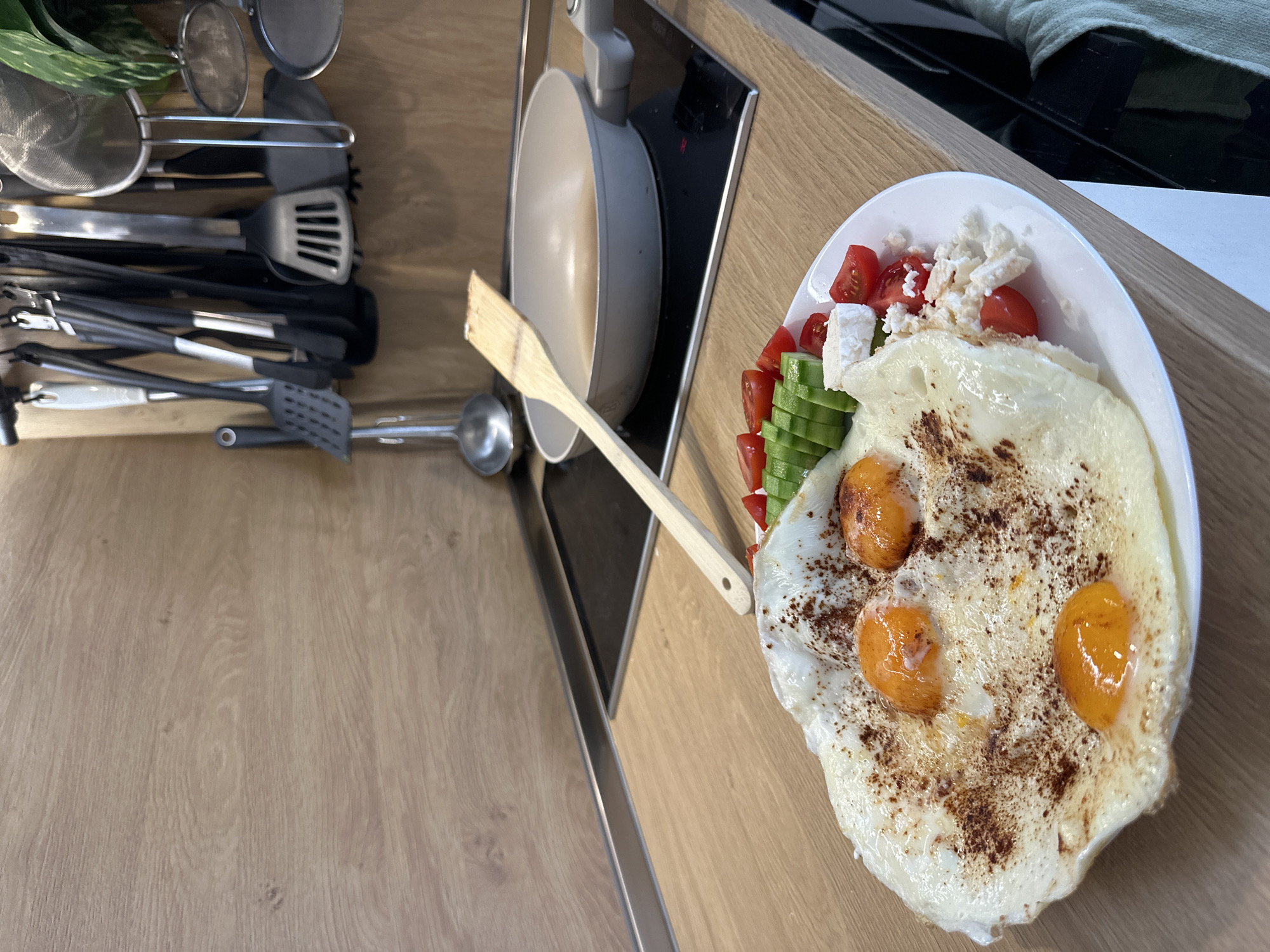 The image displays a close-up, slightly elevated view of a freshly prepared meal and kitchen area. The main subject is a white plate holding three sunny-side-up fried eggs, lightly seasoned with dark specks, possibly pepper or paprika. Accompanying the eggs are diced red tomatoes, sliced green cucumbers, and crumbled white cheese, likely feta.
The meal is presented on a light-colored wooden countertop, which features a prominent wood grain. Adjacent to the plate, a light grey non-stick frying pan rests on a sleek black induction cooktop. A wooden spoon or spatula leans against the plate, bridging towards the pan, indicating recent cooking. An "H" is visible on the cooktop's display, suggesting a hot zone.
To the upper left of the image, a collection of kitchen utensils, including various spatulas, tongs, a whisk, a strainer, and a ladle, are neatly arranged in a holder. A small green leafy plant is also visible in the top-left corner. The background on the right reveals glimpses of a dark appliance and a light green cloth.
The scene portrays a quiet, domestic setting, most likely a kitchen, during the daytime. The meal suggests breakfast or brunch. There are no people visible, and no specific location cues or visible text, aside from the "H" on the cooktop, are discernible.