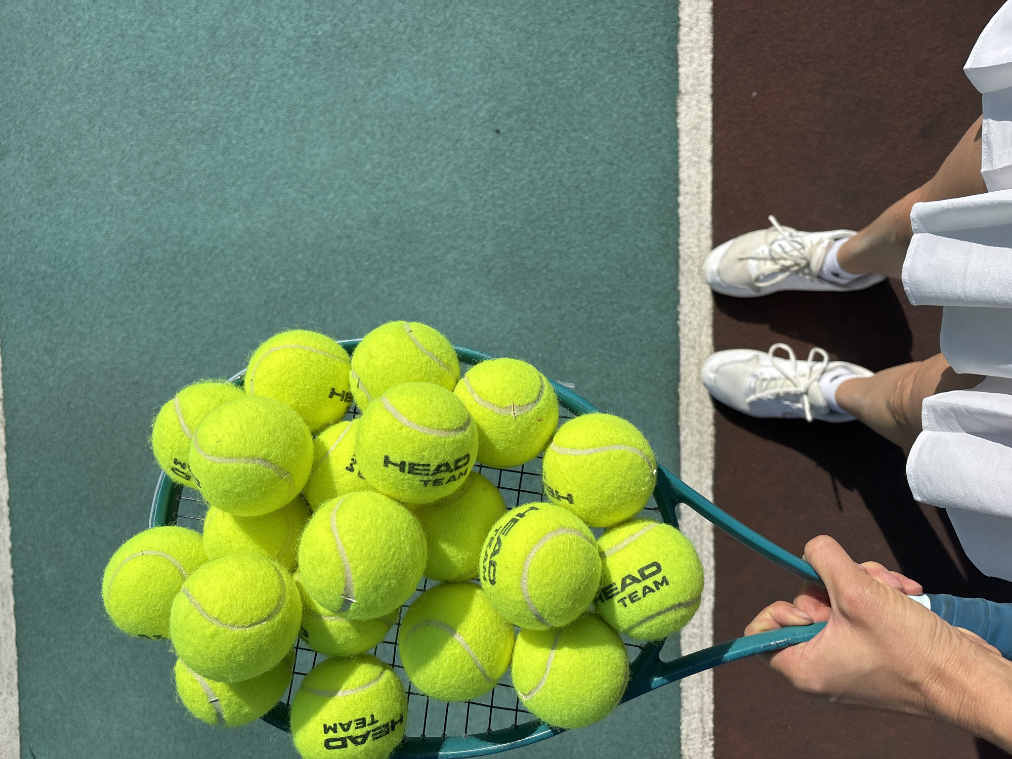A first-person shot displays a green tennis racket filled with yellow tennis balls. The tennis balls are branded "HEAD TEAM." The racket is held by two hands. The background shows a tennis court with both green and brown surfaces. In the upper right corner, the feet and legs of the person holding the racket are visible. They wear white tennis shoes and a white skirt. The exact location in Chaikovsky, Russia cannot be determined with the given image information.