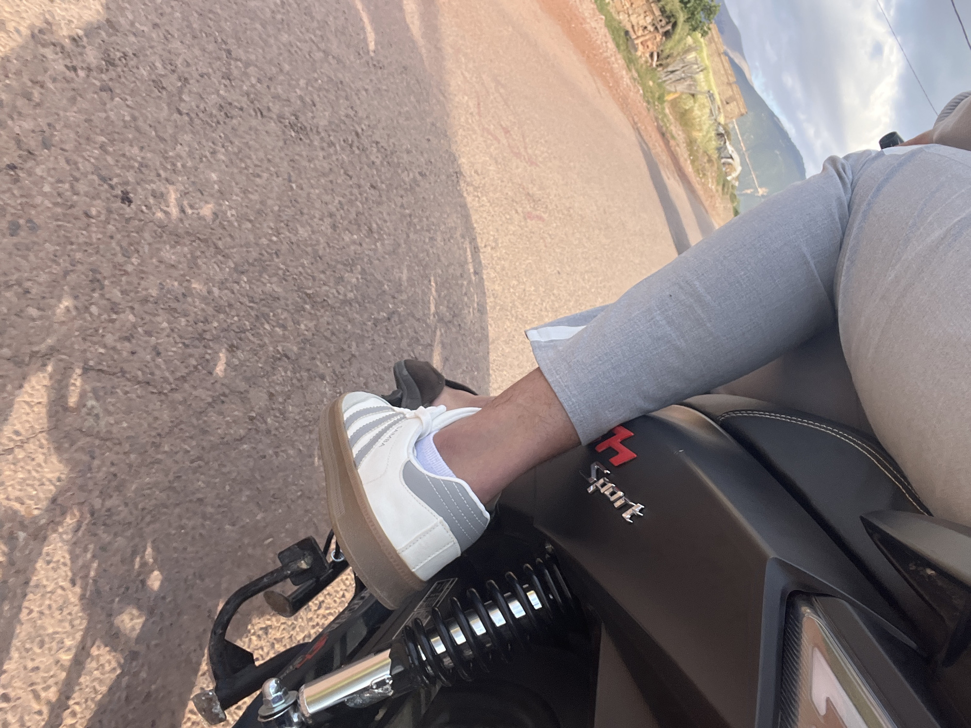 A medium shot shows a person sitting on a black motorcycle with the word "Sport" printed on its side, in what appears to be Ouaouizert, Morocco. The person is wearing light grey pants and a white sneaker with grey stripes and a brown sole. The motorcycle is positioned on a paved road with scenery showing vegetation and hills in the background under an overcast sky.
