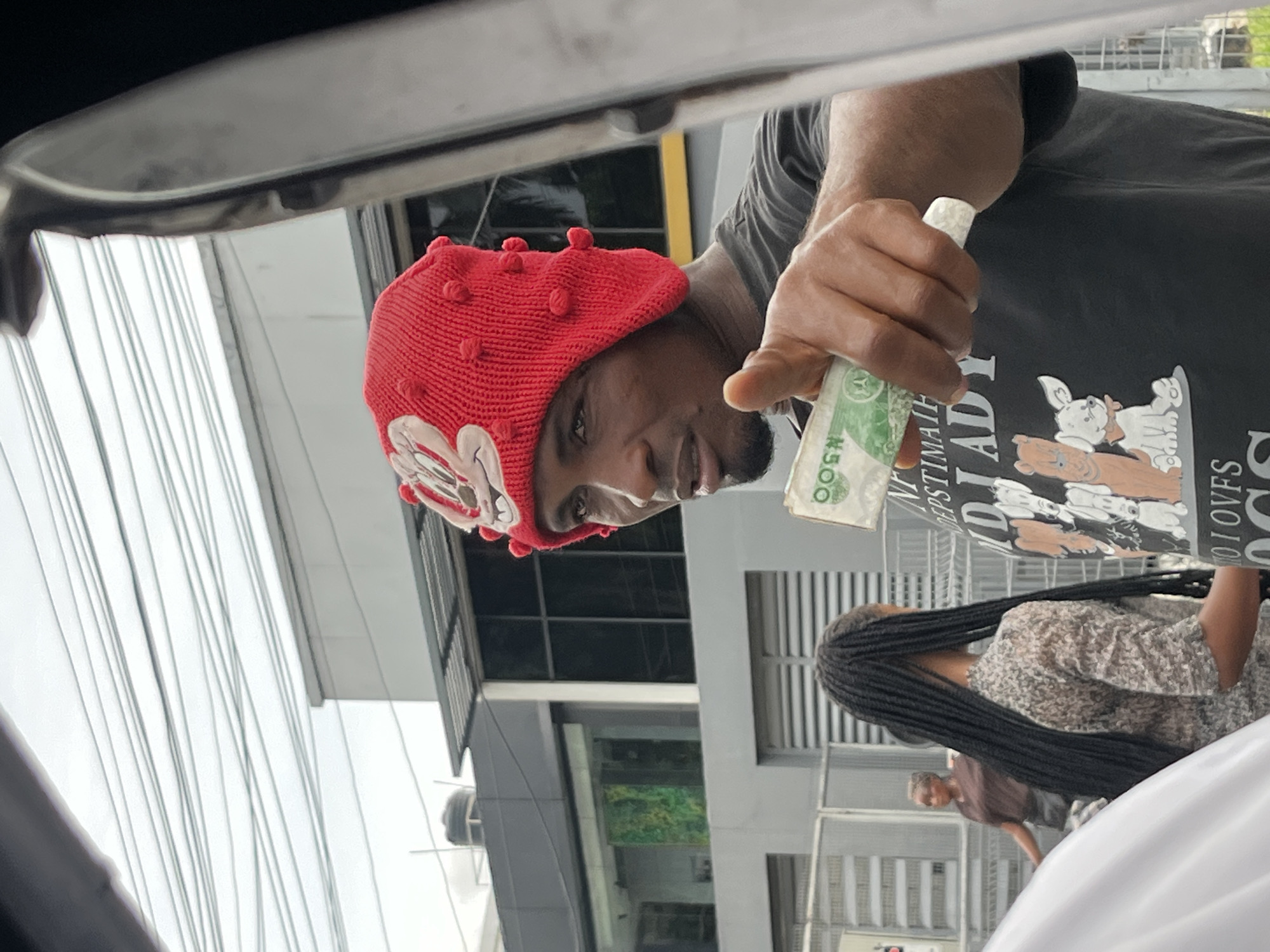 A man wearing a red knitted hat and a black T-shirt with a dog print is seen holding a wad of Nigerian Naira currency. He appears to be interacting with someone in a vehicle, judging by the car's interior visible in the frame. Another person with braided hair is standing nearby. The scene takes place in an urban environment with modern buildings and metal grid-like structures, possibly near commercial establishments. Based on the prompt, this location is identified as Itirin, Nigeria.