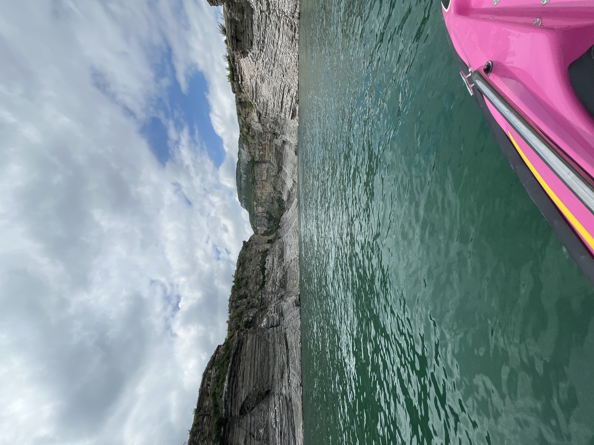 The image depicts a scene in Chirkey, Russia, showing a body of water with rocky cliffs on one side and a pink boat on the other. The water appears calm, with visible ripples and a green hue. The cliffs are gray and layered, with some vegetation growing on them. The sky is cloudy with patches of blue. A section of a pink boat is visible in the upper right corner of the frame.