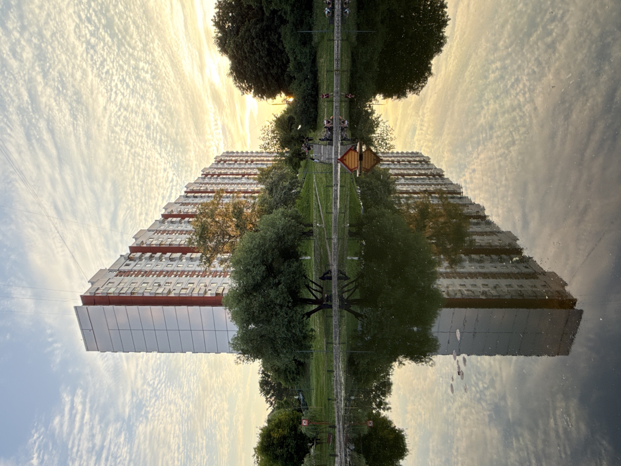 This is an upside-down, mirrored image of a cityscape at sunset, reflected in a body of water. The primary subjects are a large, multi-story apartment building with a red-accented facade and a lush green park with mature trees. The building appears to be of Soviet-era architecture, characteristic of many residential areas in Moscow, Russia.
The reflection creates a symmetrical composition, with the sky and clouds above mirroring the building and trees below. The sky is filled with wispy, textured clouds, illuminated by the warm, golden light of the setting sun, suggesting it is late afternoon or early evening. The water's surface is calm, creating a clear reflection with only minor ripples.
In the reflected scene, a pathway runs through the park, lined with trees and grass. There are a few small, red and white diamond-shaped signs on posts along the path. On the pathway, several people appear to be present, some riding bicycles or scooters and others walking. Their figures are small and indistinct due to the distance and reflection, but their presence adds a sense of life to the scene.
The overall mood is peaceful and serene, enhanced by the soft, warm lighting and the symmetrical beauty of the reflection. The location cues, such as the architectural style of the building and the presence of trees in a park setting, align with common urban landscapes found in Moscow. There is no visible text in the image.