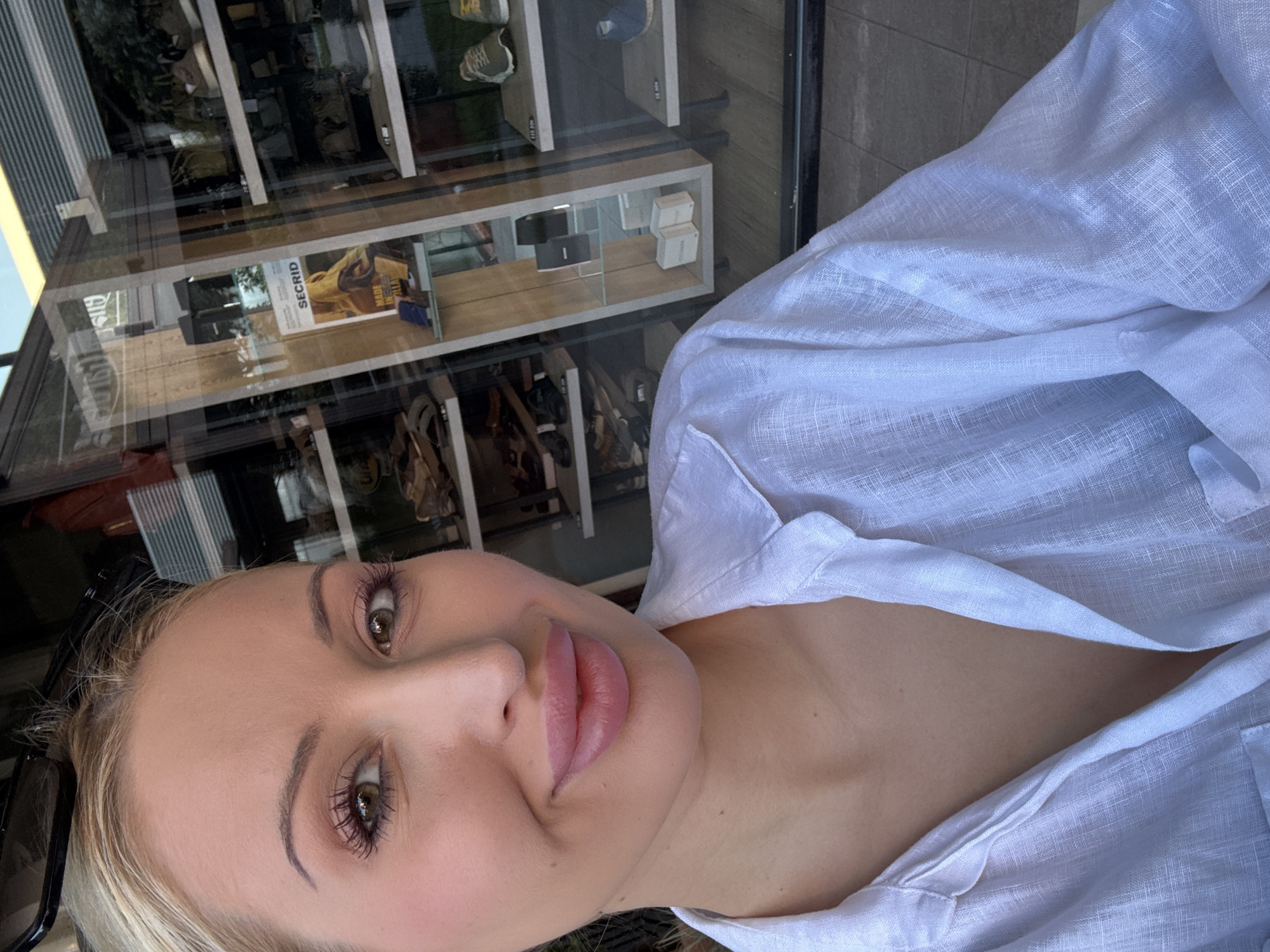 A close-up selfie shows a fair-skinned woman with blonde hair and full lips, wearing a white linen shirt. She is looking at the camera, with her mouth slightly open in a smile. In the background, a shop window displays various items, including shoes and bags, with reflections of the outdoors visible. The overall setting appears to be an urban environment, possibly a street or sidewalk.