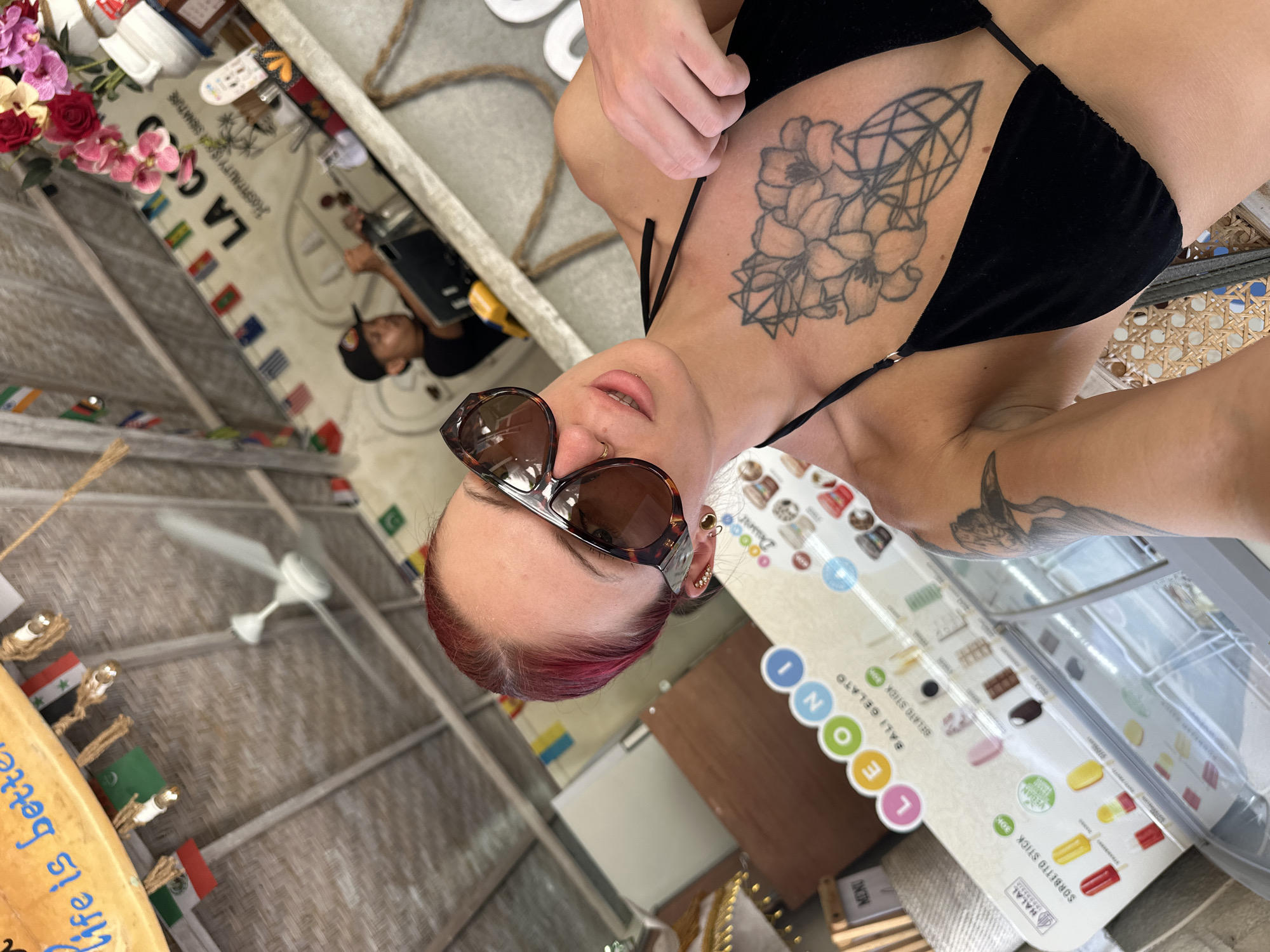 A woman with red-tinted hair and sunglasses is positioned in front of a gelato menu at what seems to be a cafe. Her visible tattoos on her upper chest and arm are notable. In the background, a sign reads "LA C," along with the phrase "HOSPITALITY IS A SIGNATURE." Decorations include international flags. The cafe appears to be in Nusa Penida, Indonesia, based on the "BALI GELATO" wording on the menu. A ceiling fan is visible.