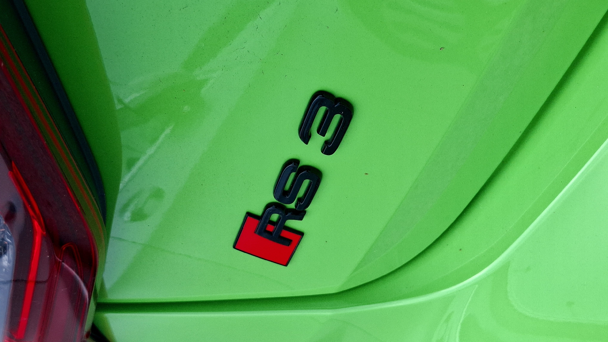 The image is a close-up, vertical shot of the rear quarter panel of a bright green car. Prominently displayed on the panel is a black and red "RS 3" badge. The "RS" has a red square behind it, and the "3" is a standalone black numeral. The car's red taillight is partially visible at the top of the frame. The green paint of the car shows a textured surface and some dust or light debris. The lighting suggests an outdoor setting, possibly a parking lot or street. The specific location of Grafenegg, Austria, is not visually represented in the image itself.