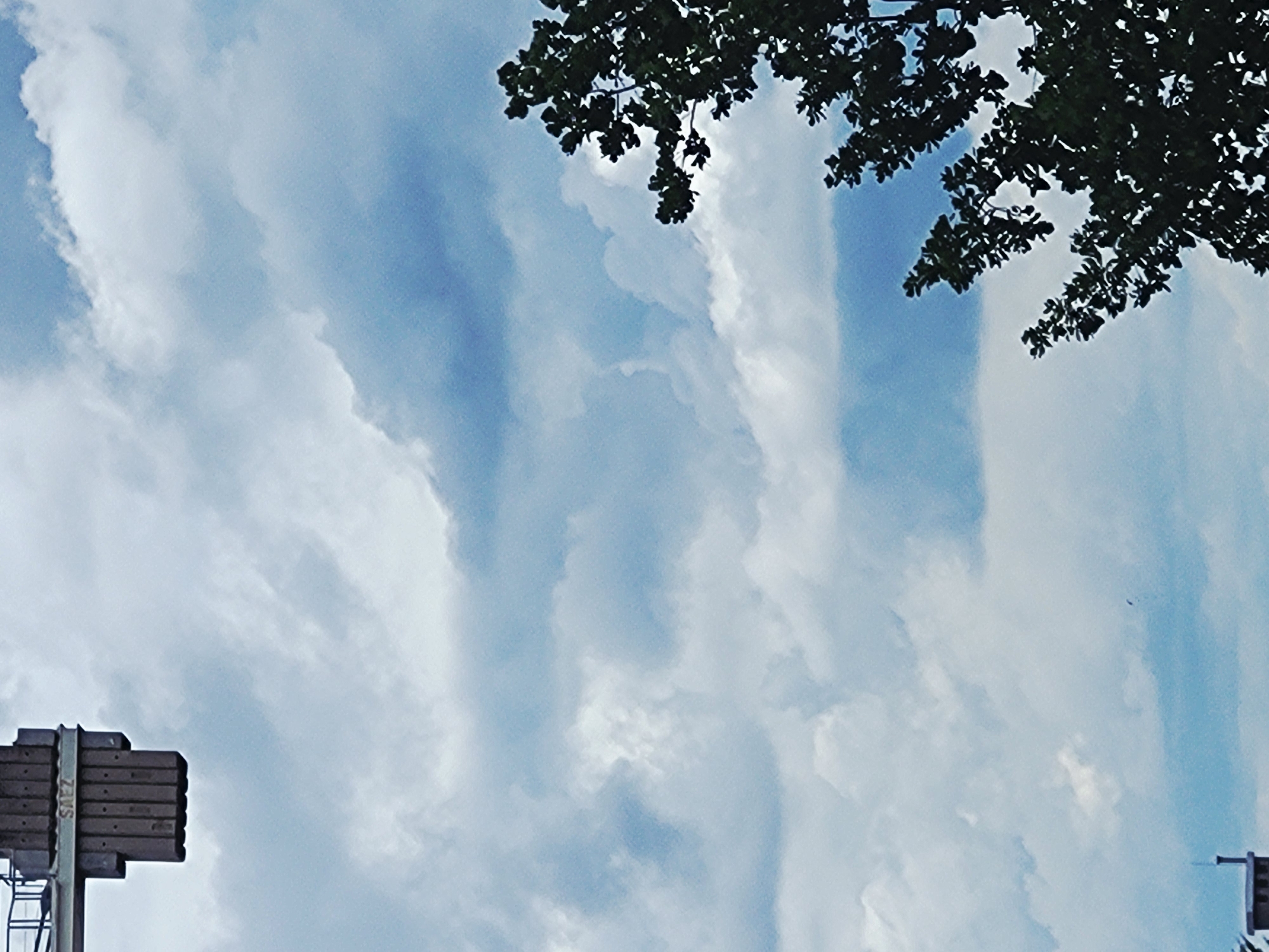 The image shows a cloudy sky with a portion of a crane in the upper left corner. The crane has a section labeled "SAEZ". In the lower right, a silhouetted tree with dark green foliage is visible against the sky. The sky is a mix of light and dark blue with white and gray clouds. A small dark object, possibly a bird or a kite, is visible in the distance against the sky. At the bottom left, a small portion of a building with a vertical structure is visible. The scene appears to be outdoors in an urban or industrial environment.