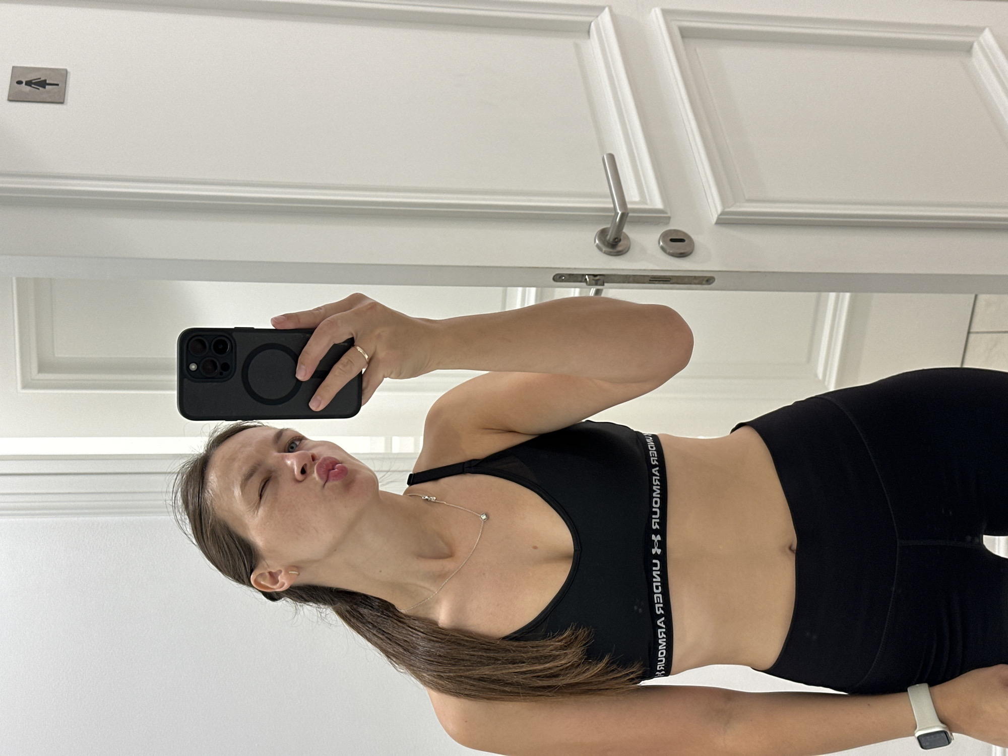 In the city of Boreti, Montenegro, a young woman is captured in a full-length mirror selfie taken in a brightly lit, modern interior space, possibly a bathroom or dressing room. She is wearing black athletic apparel, a sports bra and leggings, and her dark brown hair is pulled back into a ponytail. Her left eye is winked shut, and her lips are pursed in a playful expression. She is holding up a smartphone, which displays a black case with multiple camera lenses. A delicate necklace adorns her neck, and a ring is visible on her right ring finger. On her left wrist, a white smartwatch is worn. The background reveals a white door with classic molding, a silver doorknob, and a small, dark sign depicting a stylized female figure, indicating a women's restroom. The overall ambiance suggests a moment of personal preparation or a lighthearted selfie.
