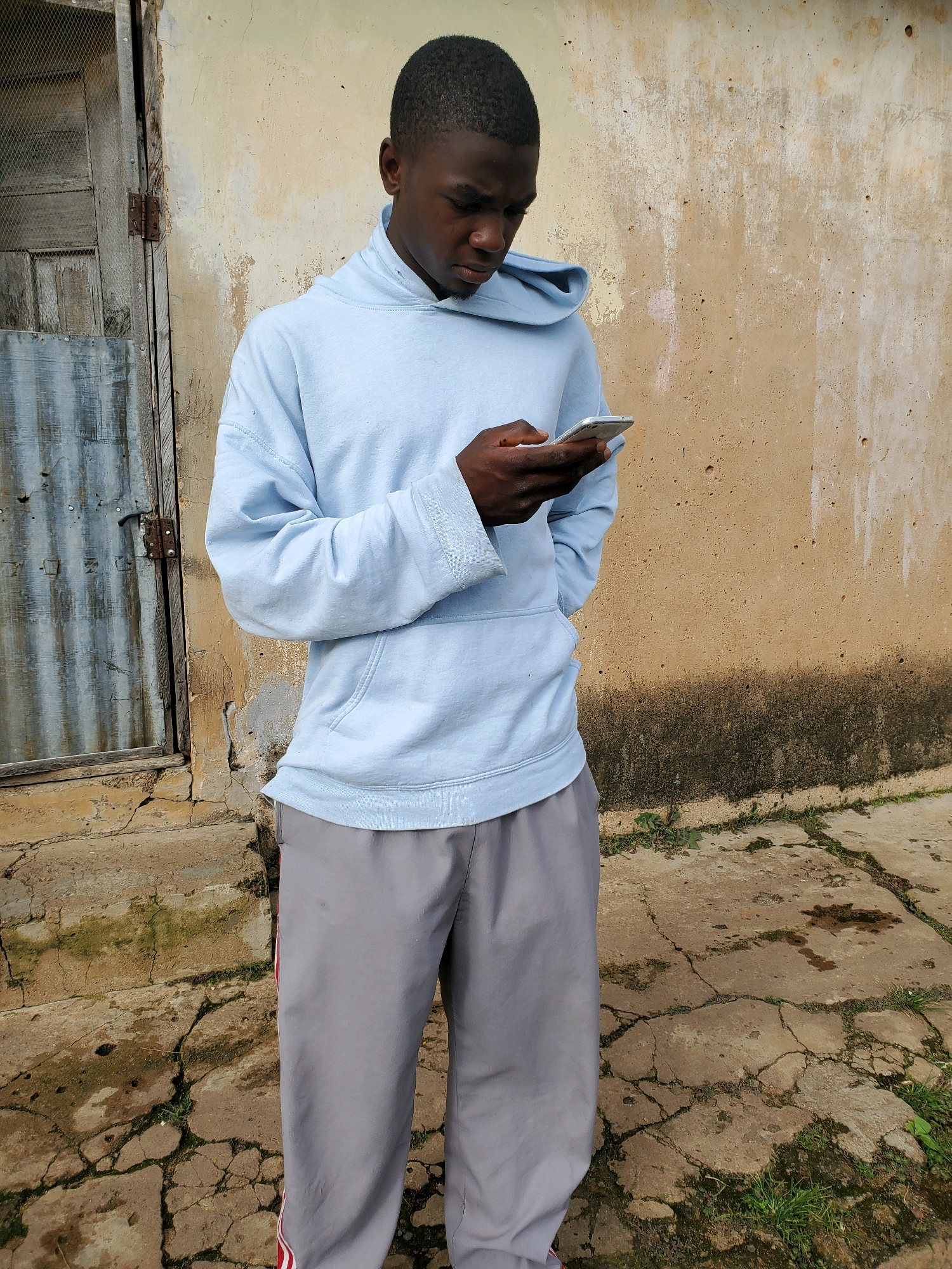 A full-body, slightly high-angle, outdoor, natural-light photograph depicts a young Black male standing in front of a weathered wall and a corrugated metal door. The subject is wearing a light blue hooded sweatshirt with a pouch pocket and grey athletic pants with a red stripe. He is looking down and holding a silver smartphone in his hands, appearing to be engaged with it. The background consists of a textured, light-colored wall with dark stains and patches of moss, suggesting an exterior surface. To the left, a rustic door with visible hinges and a metal grate is partly visible. The ground beneath is a cracked concrete surface with some patches of dry grass and darker, wet-looking areas. The scene appears to be in an outdoor urban or semi-urban environment, potentially in Jos, Nigeria, given the architectural and environmental cues.