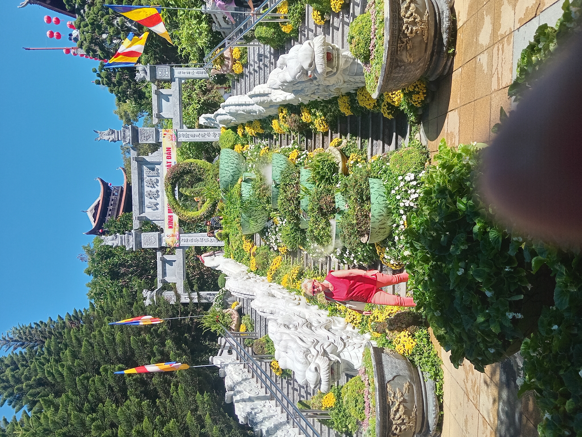 The image captures a vibrant scene at the Marble Mountains in Đà Nẵng, Vietnam. A woman in a red top and coral pants stands on a paved pathway, looking towards the camera. She is positioned in front of a grand stone staircase adorned with elaborate dragon sculptures on either side. The staircase is surrounded by lush greenery and abundant yellow and white flowers, cascading down in a picturesque display. Behind the staircase, a large, ornate stone archway stands, decorated with intricate carvings and traditional Vietnamese architecture. Two flags, one with red, yellow, and white stripes and the other with red and yellow stripes, flutter in the breeze to the left of the archway. Red lanterns are visible hanging above the archway, adding to the festive atmosphere. The environment is bathed in bright sunlight, casting shadows on the ground.