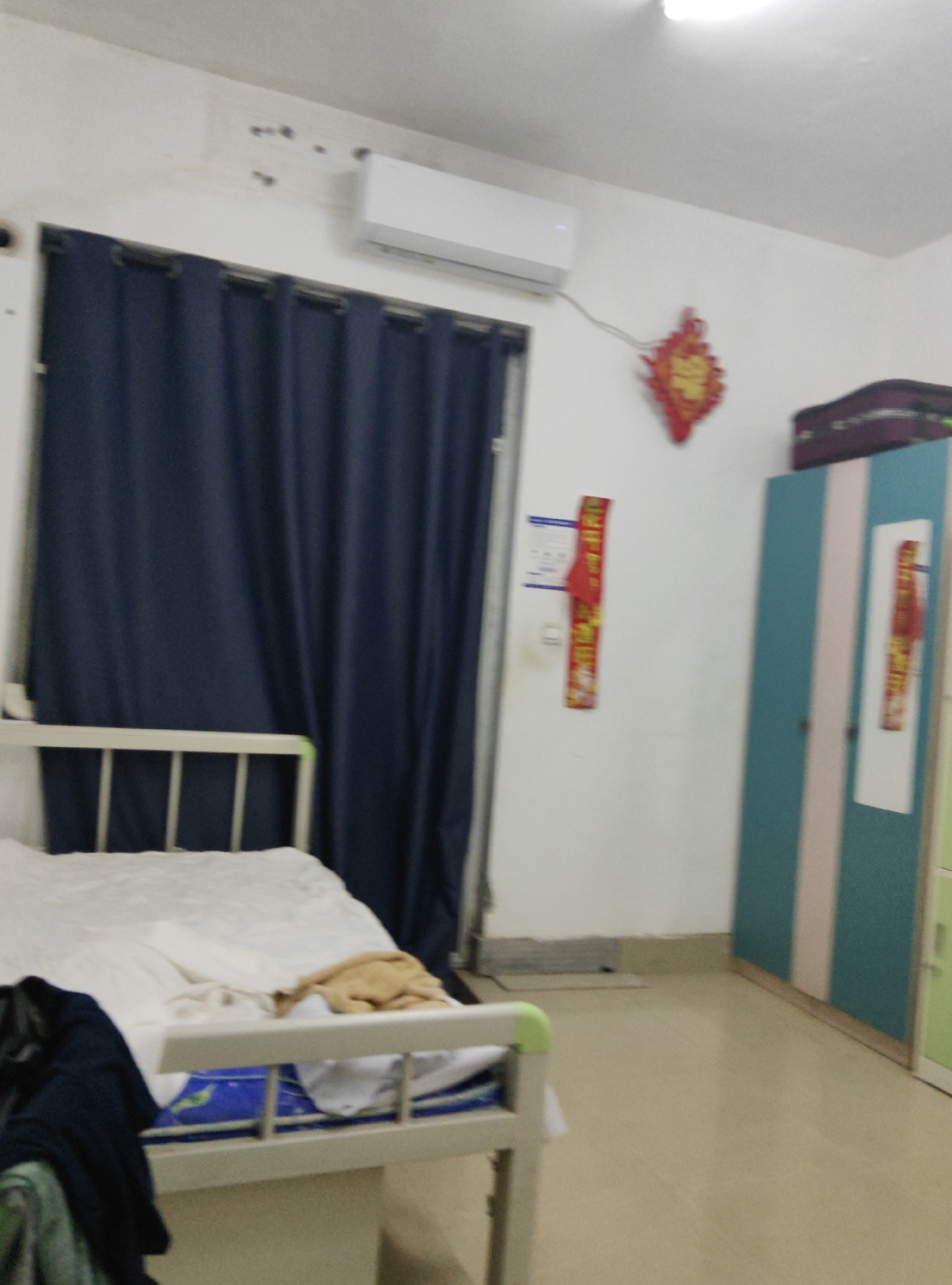 The image depicts a dimly lit, sparsely furnished room, likely a dormitory or a modest living space, located in the Chongqing High-tech Zone, China. A white-framed bed with white linens and a brown blanket is positioned in the foreground, draped with dark clothing. To the left of the bed, a dark blue curtain obscures a window. On the wall to the right, a two-toned cabinet, featuring teal and pale pink sections, stands closed. Above the cabinet, a dark suitcase rests on top. A decorative red and gold ornament, possibly a Chinese knot, hangs on the wall. An air conditioning unit is mounted high on the wall to the left, above the curtain. Another piece of red decorative paper, with Chinese characters, is affixed to the wall to the right of the air conditioner. The floor is a neutral-colored linoleum. The overall impression is one of functional simplicity, common in student accommodations or budget housing.