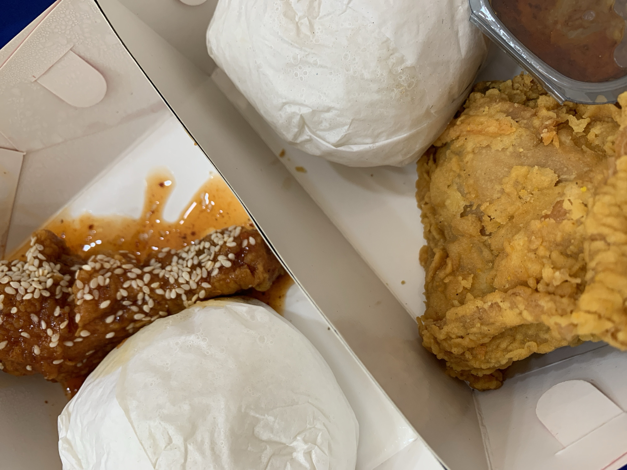 The image shows a close-up of a fast-food takeout box containing several food items. On the top left, there's a piece of fried chicken coated in a glossy, orange-brown sauce and sprinkled with sesame seeds. Next to it, a white paper wrapper encloses a round, fluffy item, possibly rice or a bun. In the bottom right section of the box, another piece of golden-brown fried chicken is visible, with a slightly uneven, crispy texture. Partially seen in the bottom right corner is a small, clear plastic container with a dark brown sauce, likely gravy or a dipping sauce. Two white paper-wrapped items are also present, one to the left of the sauce container and another to the left of the fried chicken in the top section. The box is made of white cardboard with subtle embossed patterns and a pink tab closure visible at the top. The background is a dark blue surface, suggesting the box is placed on a table or counter. There are no people or discernible environmental features indicating a specific location in Padangjaya, Indonesia, beyond the general context of a fast-food meal.