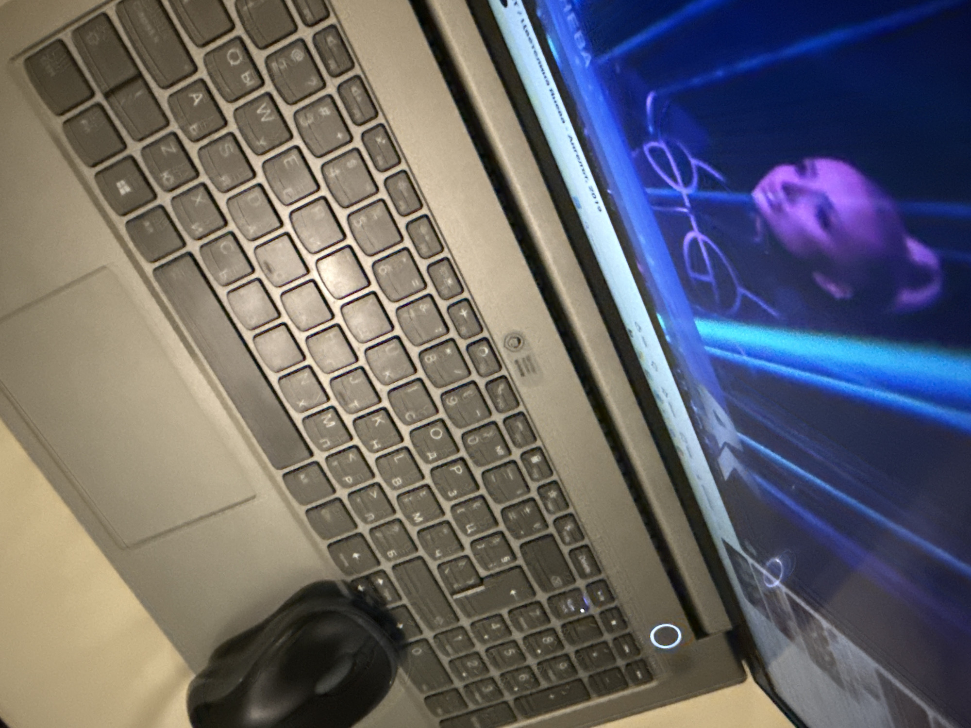 An overhead, angled shot captures a laptop on a light-colored surface, likely a desk. A black computer mouse rests near the bottom left of the keyboard. The laptop features a QWERTY keyboard with both Latin and Cyrillic characters visible on the keys, indicating a multilingual layout.
The laptop screen is illuminated with vibrant blue and purple tones, displaying what appears to be a video or image. A blurred, purple-hued human-like head or figure is discernible on the right side of the screen, seemingly surrounded by abstract, glowing lines that resemble neon or light trails.
Visible text on the screen, particularly in the top left, includes "НЕБА" and what appears to be credits or a title: "Цветелина Янева - Ангелот, 2019". "Ангелот" is a word meaning "The Angel" in some Slavic languages. The overall scene suggests a dimly lit indoor environment, possibly indicating evening or nighttime use. The activity depicted is consistent with someone using a laptop for media consumption or work. While the location context is provided as Ubud District, Indonesia, there are no specific visual cues within the image itself to corroborate this.