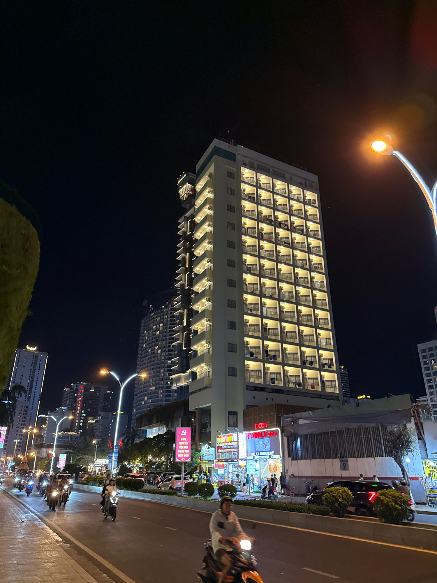 The image depicts a busy city street scene at night in Nha Trang, Vietnam. The main focal point is a tall, modern hotel building on the right side of the frame, its numerous balconies illuminated from within, creating a grid-like pattern of warm light against the dark sky. In the foreground, a wide asphalt road is filled with motorbikes and a few cars. Several individuals are seen riding motorbikes, some with passengers. To the left of the road, a paved sidewalk runs alongside the street, and beyond that, a large, green, textured structure is partially visible. Further down the street, other buildings and streetlights are visible, contributing to the vibrant, illuminated urban landscape. Signs and advertisements in Vietnamese can be seen on some of the buildings. The overall atmosphere is lively and bustling, characteristic of a commercial street at night in a Vietnamese city.