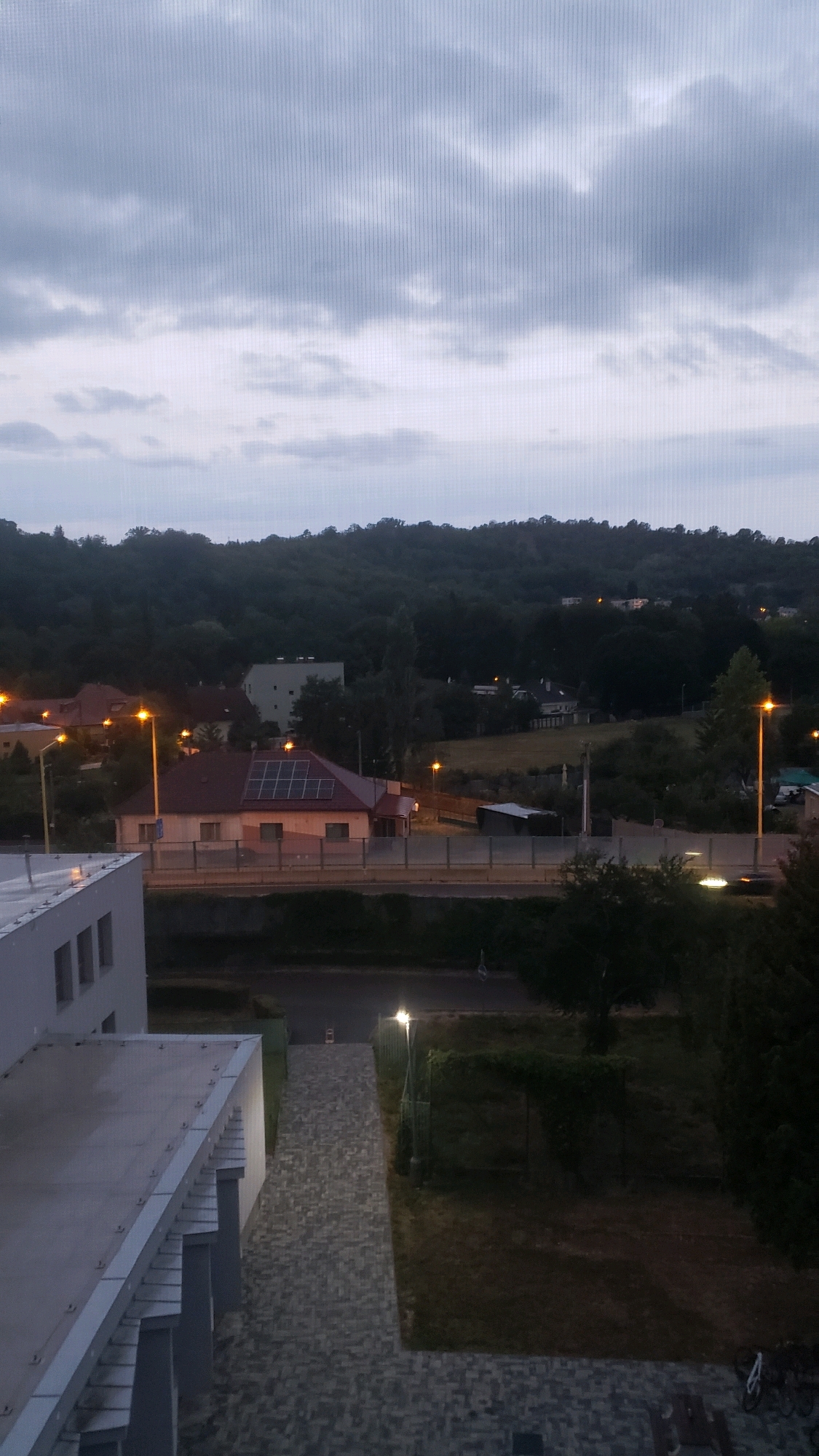 The image depicts a view from an elevated position overlooking a suburban or semi-rural area, likely at dusk. The sky is overcast with gray clouds, suggesting a dull or potentially rainy day. In the foreground, the corner of a modern, light-colored building with visible window openings is present. A flat roof with a metal edge is also visible. Adjacent to this building, a paved walkway composed of gray interlocking bricks leads away from the viewer. To the right of the walkway is a patch of mown grass, and further right is a densely vegetated area with trees and bushes.
In the midground, a street or road runs horizontally across the image. On the other side of the street, a pitched-roof house with a dark red roof and beige walls is prominent. This house has solar panels installed on its roof. Several orange streetlights are visible, illuminating the area around the house and along the street. Behind this house, a slightly larger, light-colored building with a flat roof is situated. The street is bordered by a tall, gray sound barrier or wall. Beyond the road and houses, a tree-covered hill or small mountain rises in the background, with scattered lights indicating other buildings or settlements on its slopes. The overall atmosphere is subdued and peaceful, typical of an evening in a quiet residential area.
The image does not contain any people or discernible actions taking place. The scene is static and focuses on the architectural and natural elements of the environment. Based on the provided context, this scene is located in the city of Prešov, Slovakia.