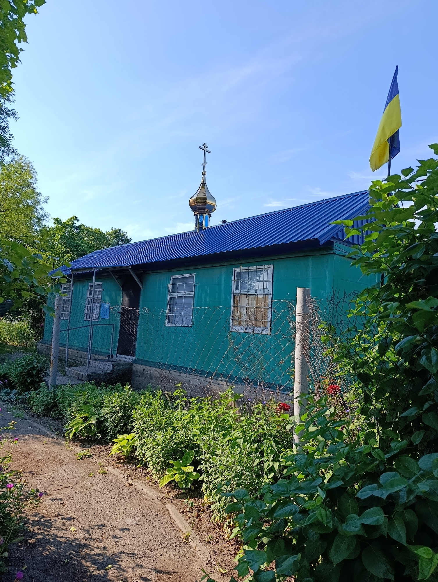 The image depicts a small, teal-colored building with a blue corrugated metal roof. A golden dome with a cross tops the structure, suggesting it is a place of worship, possibly a church or chapel. A Ukrainian flag, yellow and blue, flies from a pole on the right side of the building. The building has several windows with visible bars on the outside. A chain-link fence runs along the front of the property, separating it from a dirt path and an area with lush green foliage and some flowering plants. The scene appears to be in a rural or suburban setting, with trees visible in the background and a clear blue sky above. The overall impression is one of a modest, well-maintained religious building situated in a verdant environment.