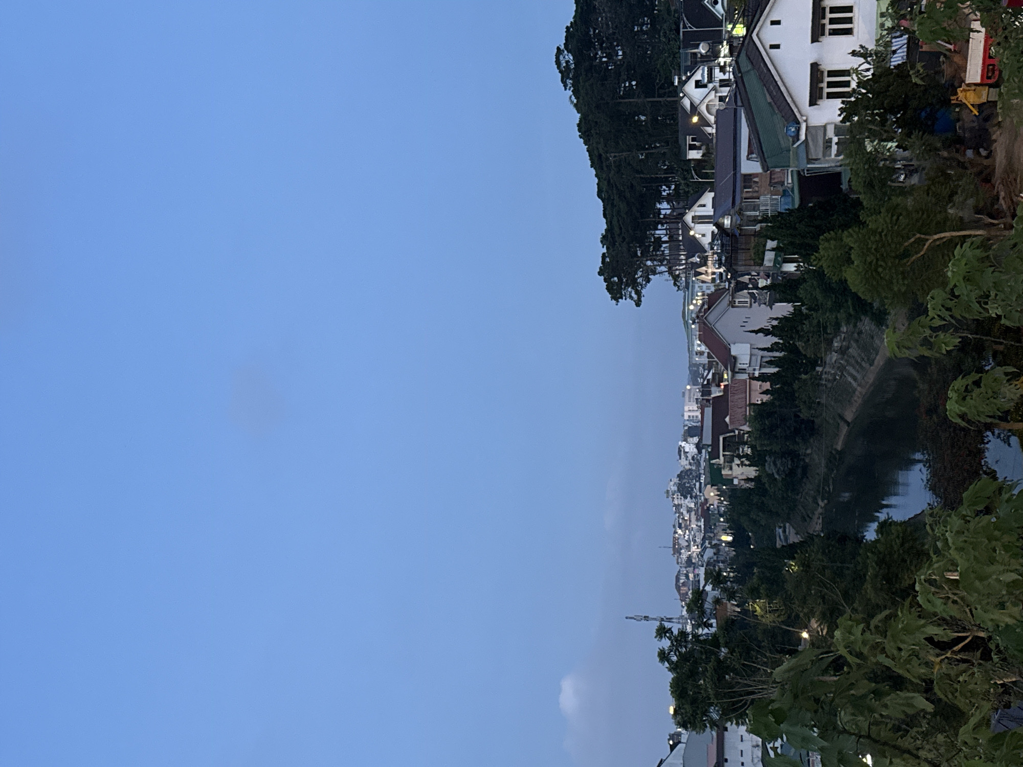 This detailed image captures a twilight or early night scene over Phường Xuân Hương - Đà Lạt, Vietnam. The upper two-thirds of the frame are dominated by a clear, deep blue sky, transitioning from dusk, with a faint, small white cloud visible on the far left horizon.
The foreground consists of dense, dark green foliage, including broad-leafed plants and trees, partially obscuring the immediate ground. A small, light-colored vehicle, possibly a utility truck or construction equipment, with a red rear section, is partially visible among the trees on the lower right side.
A dark, narrow river or canal, lined with concrete banks, meanders from the left side of the midground towards the center of the image, reflecting the low ambient light. Along both sides of the river and further into the distance, numerous residential houses are visible. These homes exhibit diverse architectural styles, with many featuring light-colored facades and various roof colors, including dark green and brown. Artificial lights are illuminated within many buildings and along what appear to be streets, creating scattered points of warmth against the cool twilight.
On the left side of the midground, beyond the river, a more densely built-up urban area extends into the distance, characterized by a multitude of buildings and numerous small lights, suggesting a city center or commercial district. A tall, slender communication or radio tower stands prominently in this left-midground section.
The right side of the midground and background is characterized by a significant natural feature: a hill densely covered with tall, dark evergreen trees, likely pines, creating a dark, textured mass against the sky. Below this tree-covered hill, more residential houses are nestled among smaller trees and shrubs. No individual people are distinctly visible in the scene.