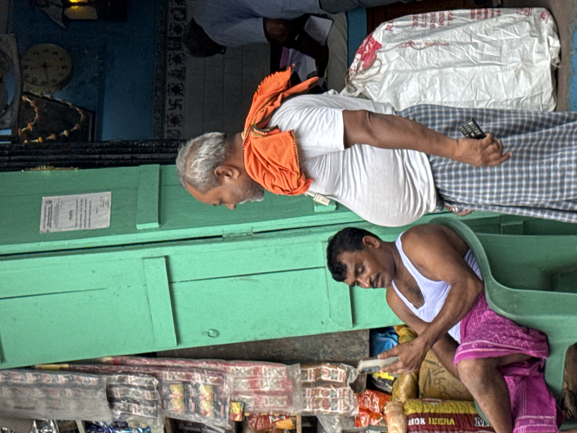 Two men in Kolkata, India are in a storefront. The man on top is older, wearing a white t-shirt, a plaid sarong, and an orange towel around his neck; he is holding a remote control. The other man is wearing a white undershirt and a pink sarong. Both men are looking down at what looks like packaged goods and snacks that are visible on shelves. The store has a green wooden counter. Visible is a wall with a clock and what appears to be swastikas. In the upper part of the image are bags, and people in the background.