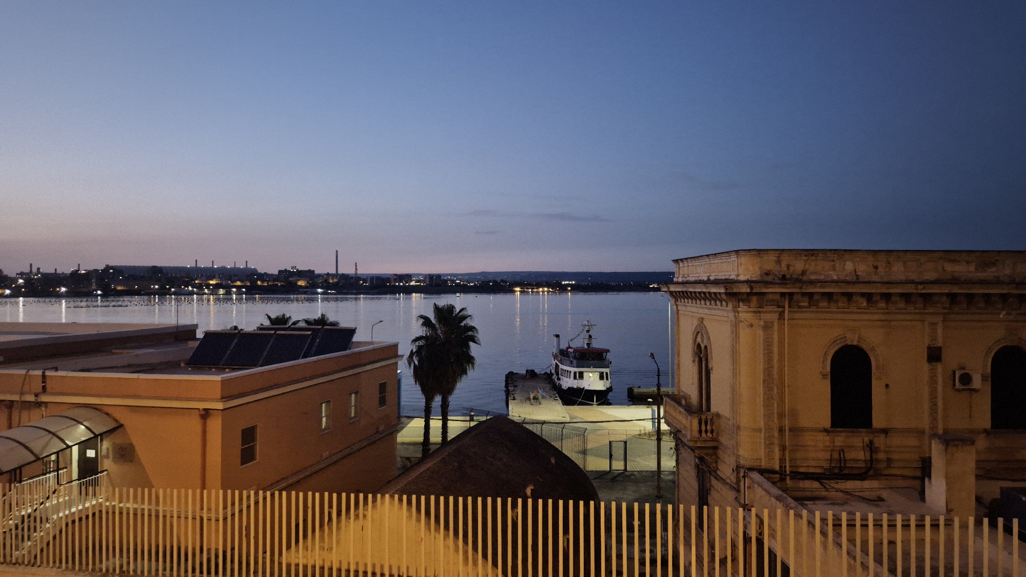 The image depicts a twilight scene in Taranto, Italy, likely overlooking a section of the Mar Piccolo or Mar Grande. In the foreground, a light-colored building with a sloped, dark-tiled roof is visible on the left, featuring a covered entryway with stairs. Adjacent to this building, a taller, light-orange building has solar panels on its roof and several windows. A white picket fence runs across the bottom of the frame, partially obscuring the lower parts of these buildings.
To the right, a more ornate, pale yellow building with arched windows and decorative architectural elements stands prominently. The middle ground reveals a harbor area with a single docked vessel, a white boat with red accents. A palm tree stands near the dock. Across the water, the distant cityscape of Taranto is illuminated with lights, suggesting an industrial or port area with multiple structures and chimneys visible against the fading sky. The overall atmosphere is calm and serene, indicative of early evening or pre-dawn twilight. There are no people or animals clearly visible in the image.