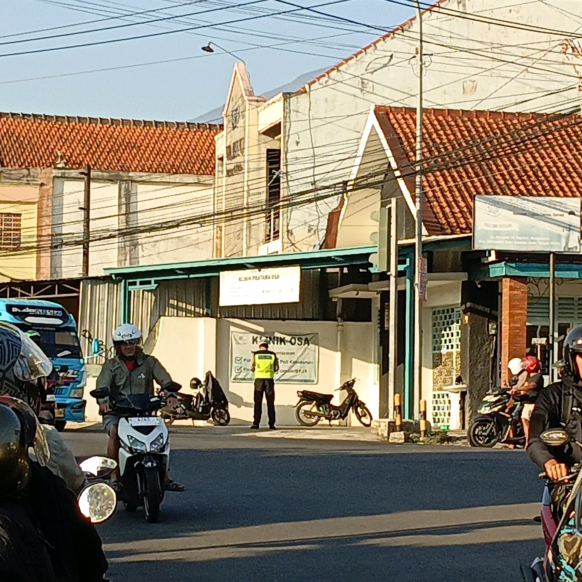 The image captures a street scene in Salatiga, Indonesia. In the foreground, a person on a white and black motorcycle, wearing a green jacket and white helmet, is stopped at an intersection. To the left, another motorcycle with a rider wearing a patterned jacket and a helmet is partially visible. Further back, a blue truck with writing on its side is parked.
In the middle ground, a person in a bright yellow vest and dark pants stands facing away from the camera, appearing to be directing traffic or performing a duty. They are positioned in front of a building with signs that read "KLINIK PRATAMA ESA" and "KLINIK OSA." Several motorcycles are parked in front of the building.
The background consists of buildings with orange tiled roofs and cream-colored walls, characteristic of Indonesian architecture. Overhead, a complex network of electrical wires crisscrosses the sky. The scene is illuminated by natural daylight, suggesting it is either morning or late afternoon.