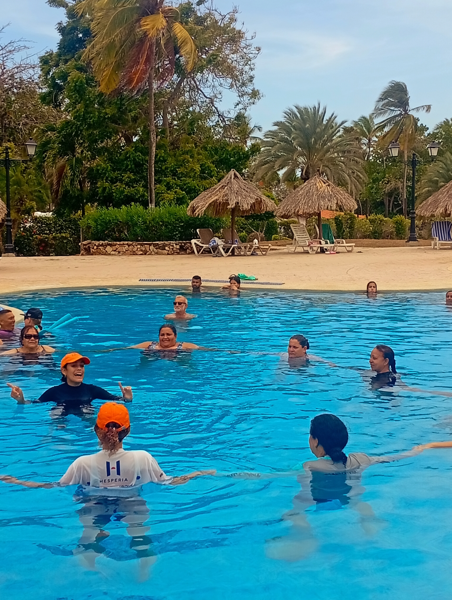 The image shows a group of people participating in a water exercise class in a swimming pool at a resort located in Playa El Agua, Venezuela. There are approximately 15 people in the pool, with two instructors leading the class. One instructor is wearing a black long-sleeved shirt and an orange baseball cap, gesturing with her hands. The other instructor is wearing a white shirt with the word "Hesperia" written on the back. The pool is surrounded by a tiled edge, and beyond that is a landscaped area with trees, palm trees, thatched-roof umbrellas, and lounge chairs. The sky is partly cloudy.