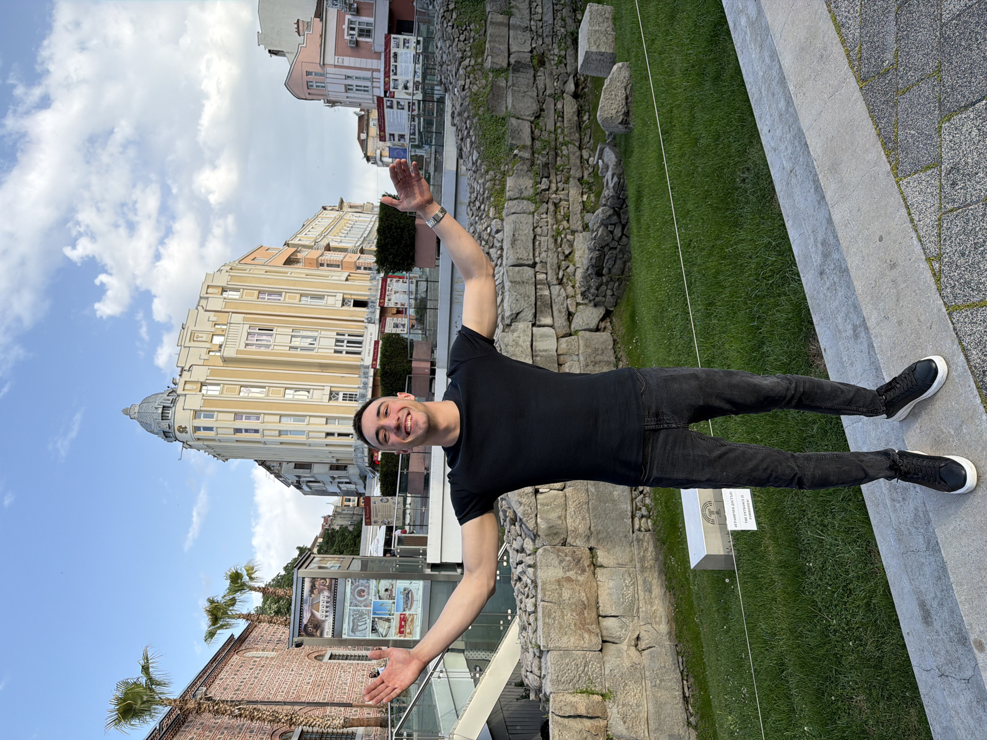 A man with dark hair and a black t-shirt is standing in an urban setting. He has his arms outstretched, and he is smiling. He is wearing black jeans and black sneakers. Behind him, there is a partially excavated ruin with stone walls. In the background, there are several buildings of varying heights and colors. To the left, there is a red brick building with palm trees in front. The sky is visible in the background with some clouds. The scene appears to be in the city of Plovdiv, Bulgaria, specifically near the Ancient Roman Theatre.