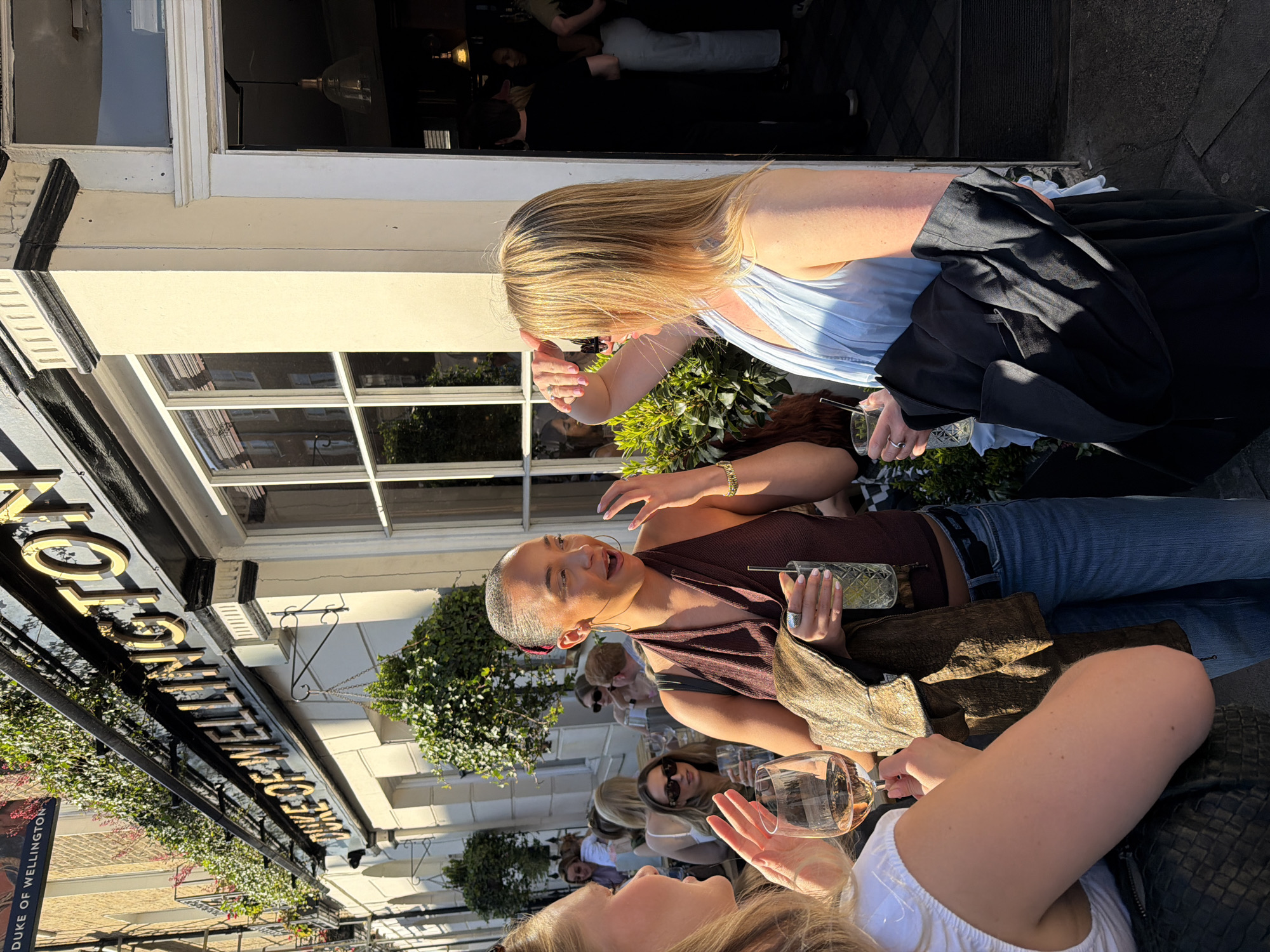 The image captures an outdoor social scene, likely in front of a pub or restaurant, identified by the prominent sign as "The Duke of Wellington." The setting is bright and sunny, suggesting a clear daytime, likely afternoon, given the strong shadows.
The main subjects are three women, with others visible in the background, all appearing to be socializing. In the foreground, on the right, a woman with blonde hair wears a light blue top, shielding her face with her hand, possibly from the sun, and holds a glass with a straw. Draped over her arm is a black jacket. To her left, another woman with dark hair, wearing a brown halter-neck top and blue jeans, gestures expressively while holding a patterned glass with a straw. She sports hoop earrings and a ring. Her expression suggests active conversation. Partially visible in the lower foreground is a third woman with blonde hair, wearing a white top and holding a wine glass filled with a light beverage, also appearing engaged in discussion.
The background shows the building facade with large windows reflecting the surroundings, and decorative green foliage in planters or hanging baskets. The overall atmosphere appears relaxed and convivial, with people enjoying drinks and conversation outdoors.
Visible text on the dark overhead sign includes "DUKE OF WELLINGTON" and "WELLINGTON" in gold lettering.