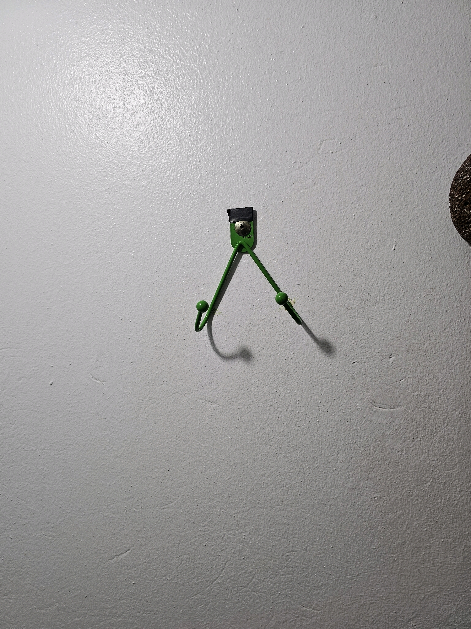 A close-up, eye-level shot captures a green double-hook mounted on a textured, light gray wall. The hooks are shaped like an inverted V, with the base attached to the wall by a screw and a black piece of tape. A small, dark, round object, possibly a decorative sphere or a part of another object, is partially visible in the upper right corner of the frame. The lighting casts a shadow of the right hook onto the wall, creating a sense of depth. The scene is devoid of any specific identifying features that would allow for pinpointing its exact location within the city of Batna, Algeria.