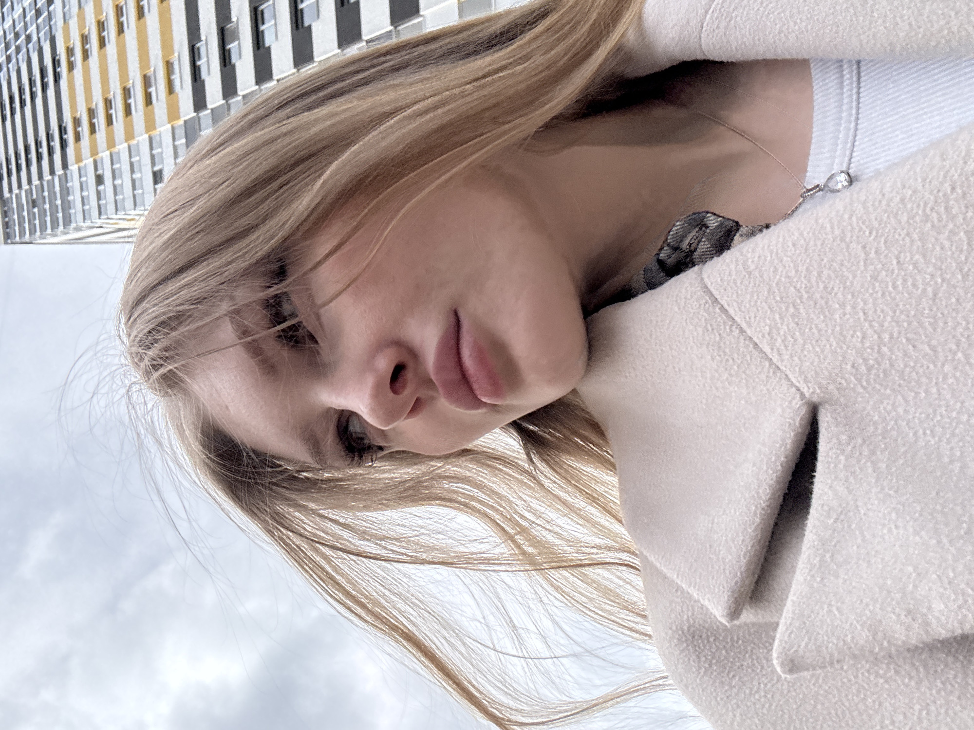 A low-angle, slightly rotated close-up captures a fair-skinned young woman with long, blonde hair. She wears a light-colored coat, revealing a white top and a thin necklace with a small pendant. The backdrop includes a tall building with alternating yellow, white, and black stripes, suggesting a modern architectural design. Above her, a cloudy sky is visible. The image location is likely Perm, Russia, based on visual context, but a specific street or landmark is unclear. No discernible interactions or activities are evident.