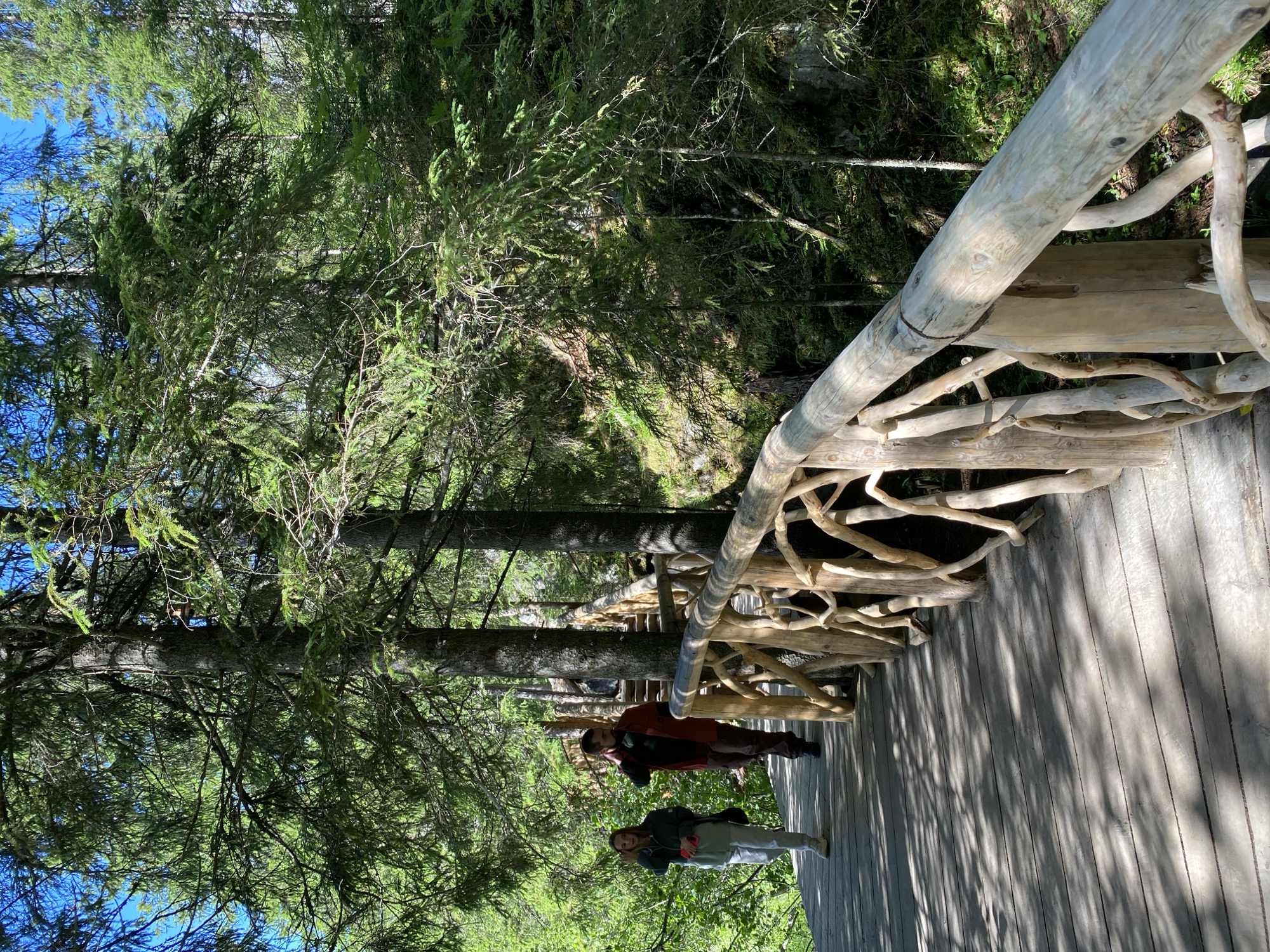 In Ruskeala, Russia, a wooden walkway winds through a dense forest with tall evergreen trees and lush green foliage. The walkway features a rustic, naturalistic railing made of intertwined branches. Two individuals are walking along the path. In the foreground, a young woman with light-colored hair, wearing a light green jacket and light pants, is seen from the waist down. She is holding a red object in her left hand. Behind her, a man wearing a reddish-orange jacket and dark pants walks away from the camera, his left hand near his neck. The sun casts dappled shadows on the wooden planks of the walkway, creating a play of light and dark. The environment suggests a natural, outdoor setting, likely a park or scenic trail.