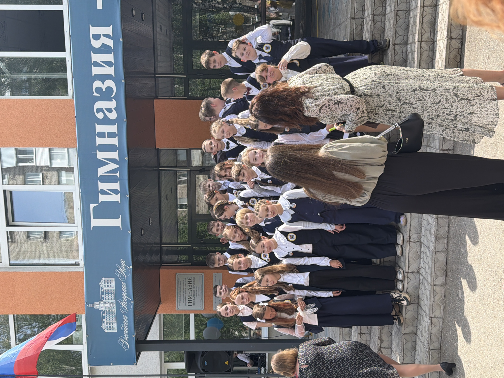 A group of approximately 30 students, mostly girls, are posing for a photograph on the steps of a building with a sign that reads "Гимназия" (Gymnasium) in Russian. The building, identified as a gymnasium in the city of Chaikovsky, Russia, has a blue banner with the Russian flag visible on the left. The students are wearing uniforms with white tops and dark skirts or pants. Several adults, including a woman in a floral dress and another in a black dress, are present and appear to be involved in organizing the photo. The location is outdoors on a sunny day.