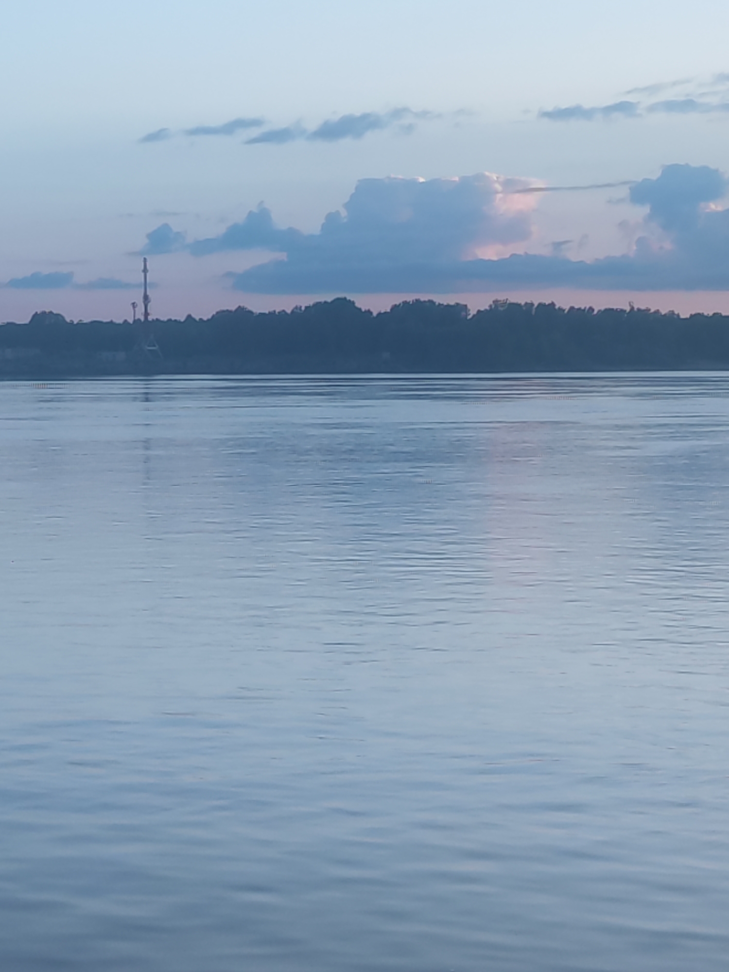 The image presents a tranquil landscape featuring a wide body of water under a pastel sky, likely captured during dawn or dusk in Noviy, Russia. The lower two-thirds of the frame are dominated by the calm water, which displays gentle ripples and reflects the subtle blue-grey and pinkish-purple hues of the sky.
A dark, indistinct silhouette of a treeline or landmass stretches across the distant horizon. On this remote bank, slightly to the left of the center, a tall, slender structure, possibly a communication tower, stands out, with its blurred reflection visible in the water.
The sky, filling the upper third of the image, features soft blue-grey tones transitioning to delicate pinkish-purple, particularly near the horizon. Scattered clouds are present, with some showing darker undersides and a distinct area of warm, pinkish-orange light breaking through a gap in the clouds on the right, indicating the sun's low position.
No specific activities or people are visible, contributing to the scene's serene and contemplative mood. The weather appears calm, with soft, diffused lighting. There is no discernible text within the image.