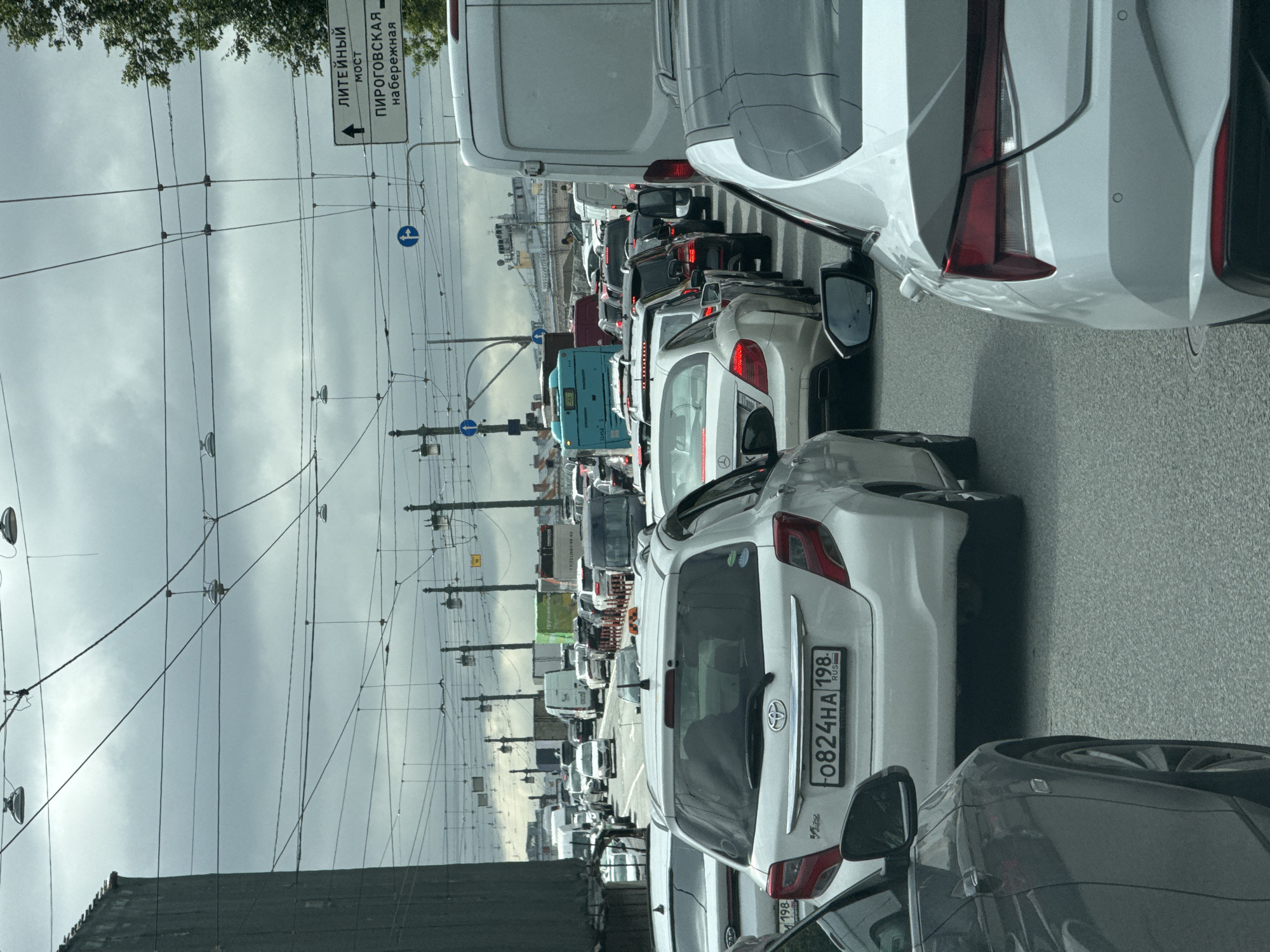 The image shows a traffic jam on a cloudy day in Saint Petersburg, Russia. Multiple lanes of vehicles are stopped on a road with overhead power lines and traffic signals. A street sign with Russian text, indicating directions to "Liteynyy Most" and "Pirogovskaya Naberezhnaya," is visible on the right. Several cars, including a white Toyota Vitz with the license plate F0824HA 198, a white Mercedes-Benz, and a dark grey car, are prominent in the foreground. A blue bus and several other vehicles are also part of the congestion. The environment suggests an urban setting with buildings and trees visible in the background.