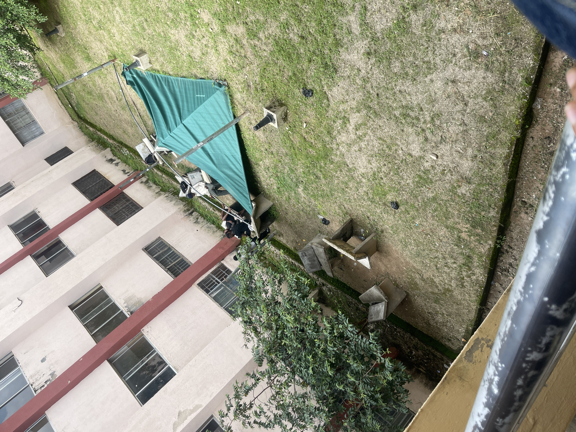 The image, taken from a high vantage point, depicts an outdoor courtyard area adjacent to a multi-story building. The building, painted a light color with prominent reddish-brown horizontal bands and windows, occupies the left side of the frame. The ground is covered in grass, with patches of dry soil.
In the center of the courtyard, a large, green tarp is supported by metal poles, suggesting a temporary shelter or awning. Beneath the tarp, several objects are visible, including what appear to be bags and possibly some sort of equipment. A person in dark clothing is partially visible near this setup, looking down towards it.
Scattered across the grassy area are several concrete structures, some appearing to be planters or perhaps part of a disused seating area. A small, leafy tree grows in the lower right portion of the courtyard, its branches reaching upwards.
The overall scene suggests a somewhat unkempt or utilitarian outdoor space. The lighting indicates it is daytime, but the sky is not visible, so the specific weather conditions are not discernible. The presence of the tarp and the objects beneath it hints at some activity taking place, possibly for shade or protection from the elements. The perspective from above and the angle of the building suggest the photo was taken from a balcony or window of an upper floor. No discernible text is visible in the image.