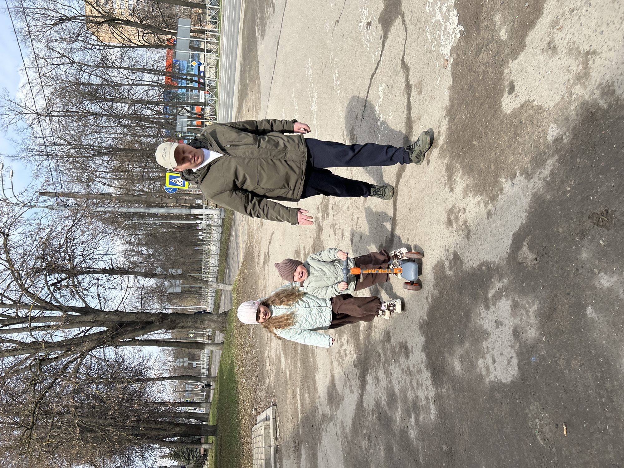 The image shows three people on a paved surface in Klin, Russia. An adult male stands in the upper portion of the image, wearing a green jacket, dark pants, green sneakers, and a white baseball cap. Two children are in the middle of the image. They are wearing matching light green quilted jackets and brown pants. One child wears a light pink beanie and the other wears a darker brown beanie. They are riding a small blue and orange toy vehicle with white shoes. The background includes bare trees lining a grassy area on the left side of the image. A building with blue, orange, and white panels is visible in the distance. A street sign is partially visible near the adult male. The ground appears to be a mix of dry and damp pavement.