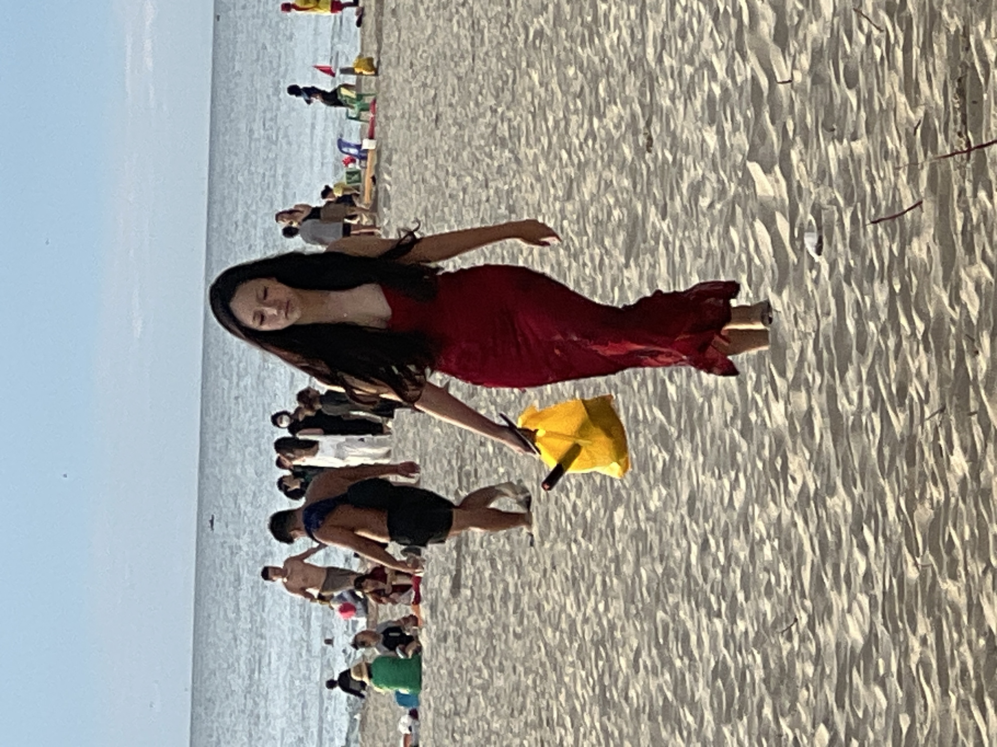 A young woman with long, dark hair, wearing a red, floor-length dress, walks on a sandy beach towards the viewer. She is holding a yellow bag in her left hand, with a dark object protruding from it. The beach is populated with other people in the background, some in swimwear, others in casual clothing. Some are sitting, others are standing and interacting. The ocean is visible behind the beach, with a calm surface under a hazy sky. The scene appears to be on a beach in Da Nang, Vietnam, given the context provided.