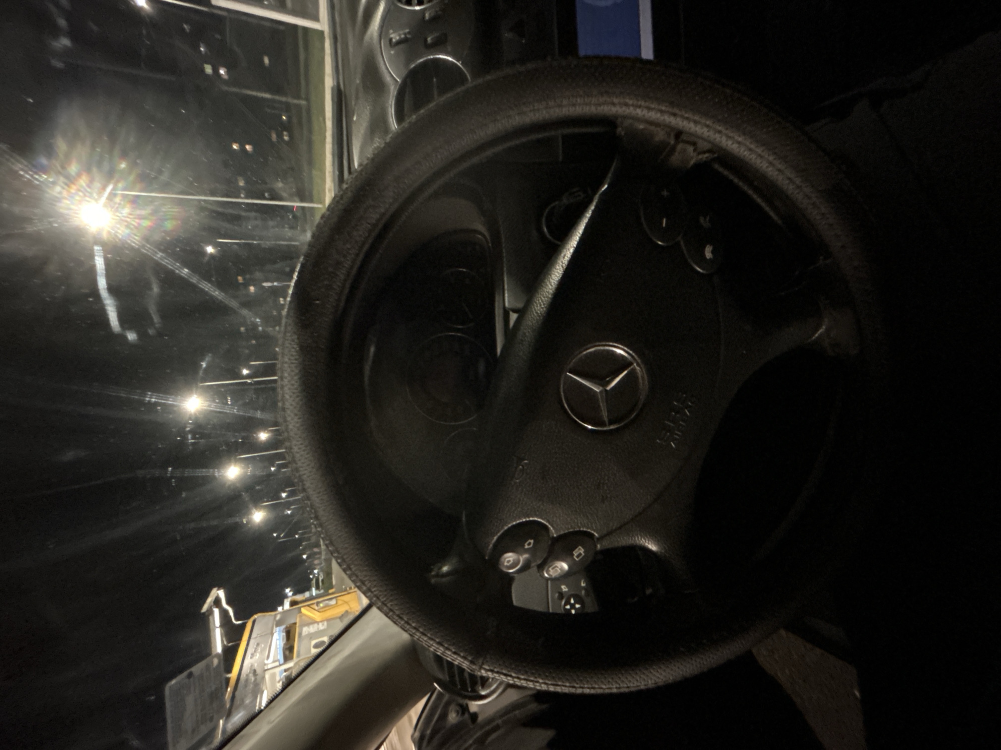 The image depicts the interior of a vehicle, specifically the steering wheel of a Mercedes-Benz car. The steering wheel is black and has a steering wheel cover on it. The Mercedes-Benz logo is prominently visible in the center of the steering wheel. Various buttons and controls are integrated into the steering wheel design. Through the windshield, the outside environment appears to be illuminated by streetlights. The presence of construction equipment or vehicles is visible, possibly from a construction site, illuminated by lights in what seems to be a dark environment. The location, Vratsa, Bulgaria, is only known from the prompt. It cannot be verified from the image itself. No people are visible, but the perspective suggests the photo was taken from the driver's seat.