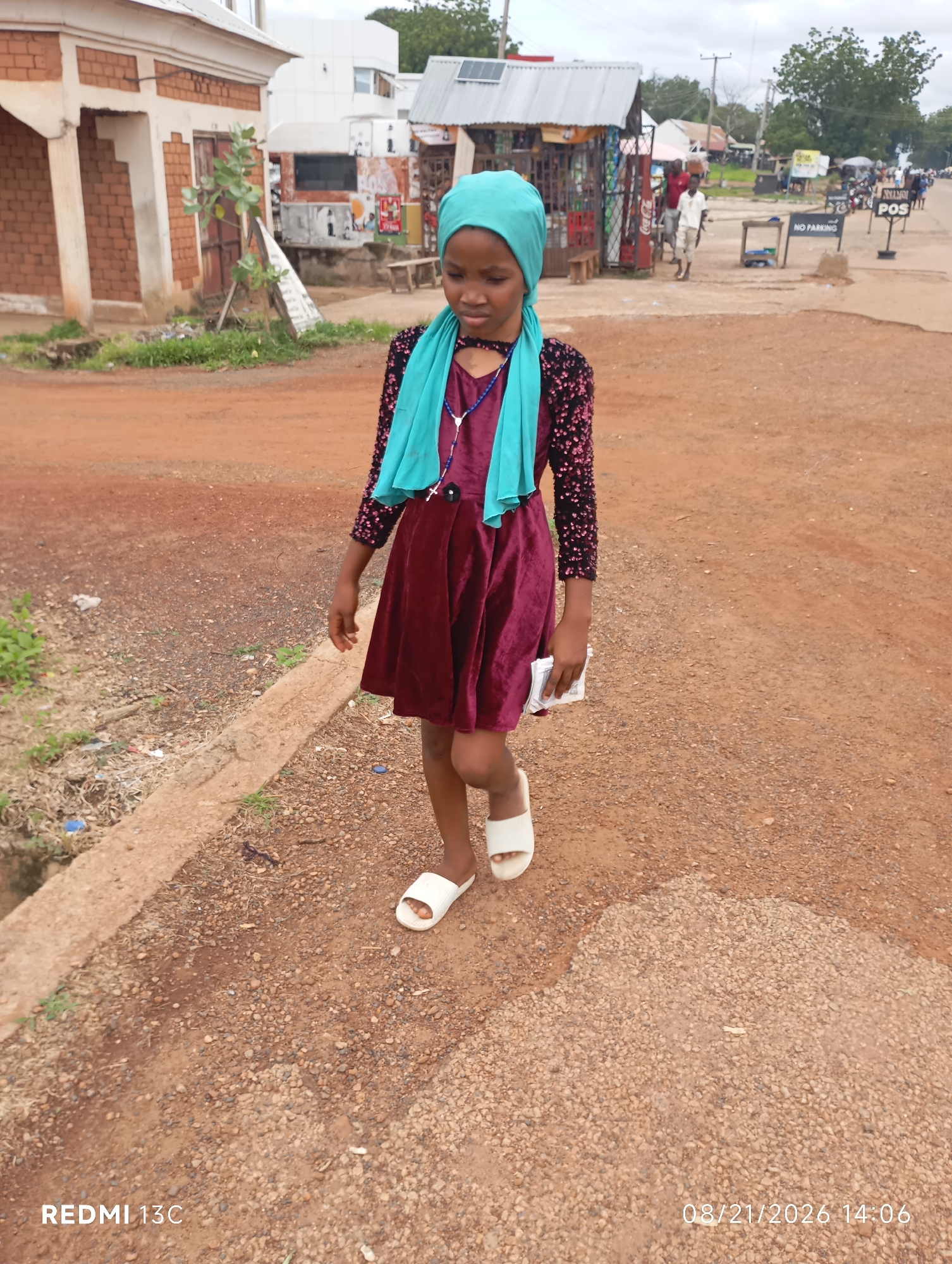 A young Black girl wearing a teal headscarf, a burgundy velvet dress with a sequined bodice, and white sandals walks on a dirt road in the city of Shendam, Nigeria. She is holding a small stack of papers in her right hand. The road is lined with a concrete curb, and beyond that, there is a small drainage ditch with some debris and vegetation. In the background, there are several buildings, including a brick structure to the left and small market stalls with various goods displayed. There are also other people visible in the distance, as well as road signs, including one that reads "NO PARKING" and another that says "POS". The sky is overcast. The timestamp on the image indicates it was taken on August 21, 2026, at 2:06 PM. The device used to capture the image is a REDMI 13C.