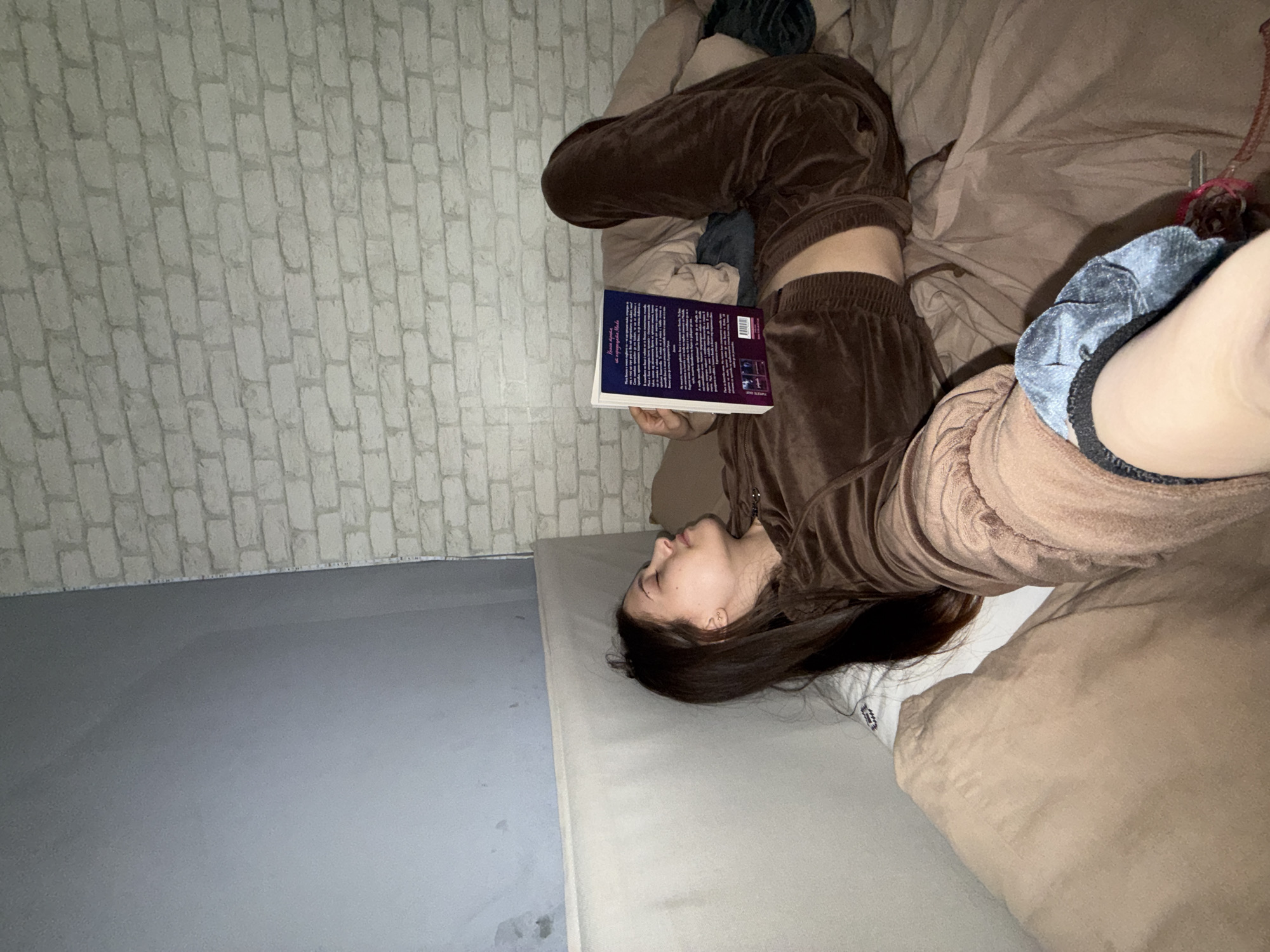 A young person, likely female, is depicted lying on their back on a bed, engaged in reading a book. They are dressed in a brown velvet tracksuit. The book has a purple spine and cover, and is held open over their torso. Visible text on the book cover includes "Последна страница" (Last Page) and "На български език" (In Bulgarian language). A dark blue velvet scrunchie is worn on their left wrist, alongside what appears to be keys with a red accessory. A white sock with the word "SPORT" is visible on their left foot.
The setting is an indoor room, likely a bedroom, characterized by a light grey wall with a distinct brick-like pattern on the left side of the frame. The lower left corner reveals a plain grey floor. The bed consists of a light-colored mattress and brown bedding. The overall lighting is dim, suggesting it is evening or nighttime. The person appears calm and focused on their reading. The location context provided is Sofia, Bulgaria.