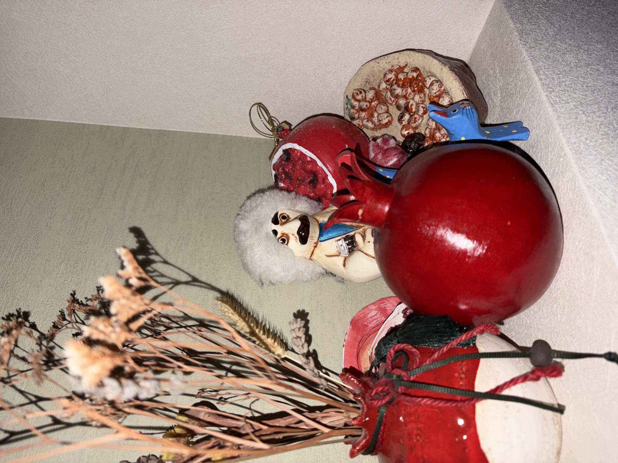 The image shows a still life arrangement on a white shelf in an interior. The arrangement includes a red vase with dried flowers, several pomegranate-themed decorations, a figurine of a man with a white fluffy hat and mustache, and a small blue cat figurine. The background consists of two walls, one light green and textured, and the other off-white and also textured. No information about the city of Chelyabinsk, Russia, or any specific location is present in the image. The scene appears to be a decorative display, possibly in a home or shop.