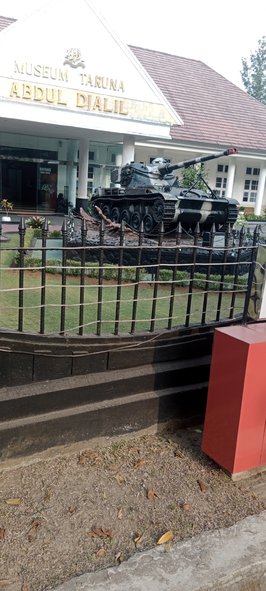 The image depicts the Museum Taruna Abdul Djalil in Magelang, Indonesia. A military tank, camouflaged with dark green and white patterns, is displayed prominently in front of the museum entrance. The museum building has a white facade with a triangular roof and large glass windows. A black metal fence with decorative spikes and horizontal ropes separates the foreground from the museum grounds, where the tank is situated. The grounds feature a manicured lawn and some shrubbery. In the lower right portion of the image, a bright red rectangular object, possibly a trash bin or display element, is visible. The foreground consists of a dark, textured surface, possibly concrete steps or a pathway, with some dried leaves scattered on the ground. The sky is overcast.